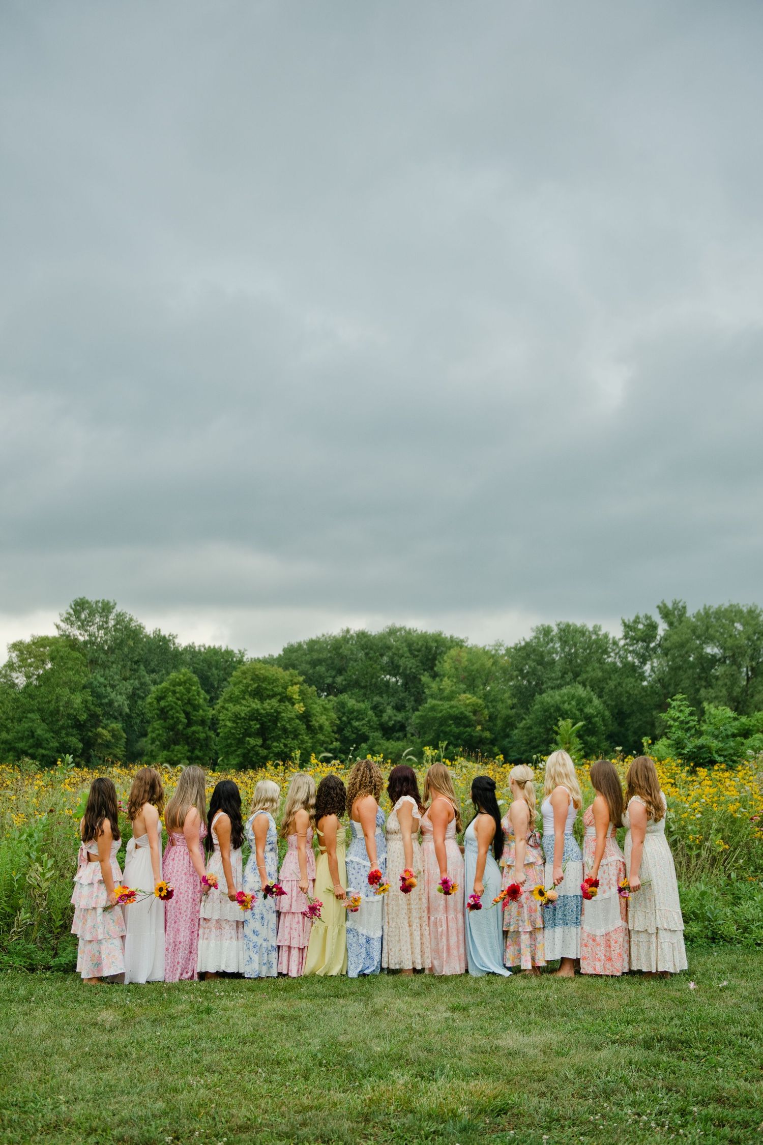 Young women in colorful pastel dresses stand with backs to camera holding flowers in a green field.