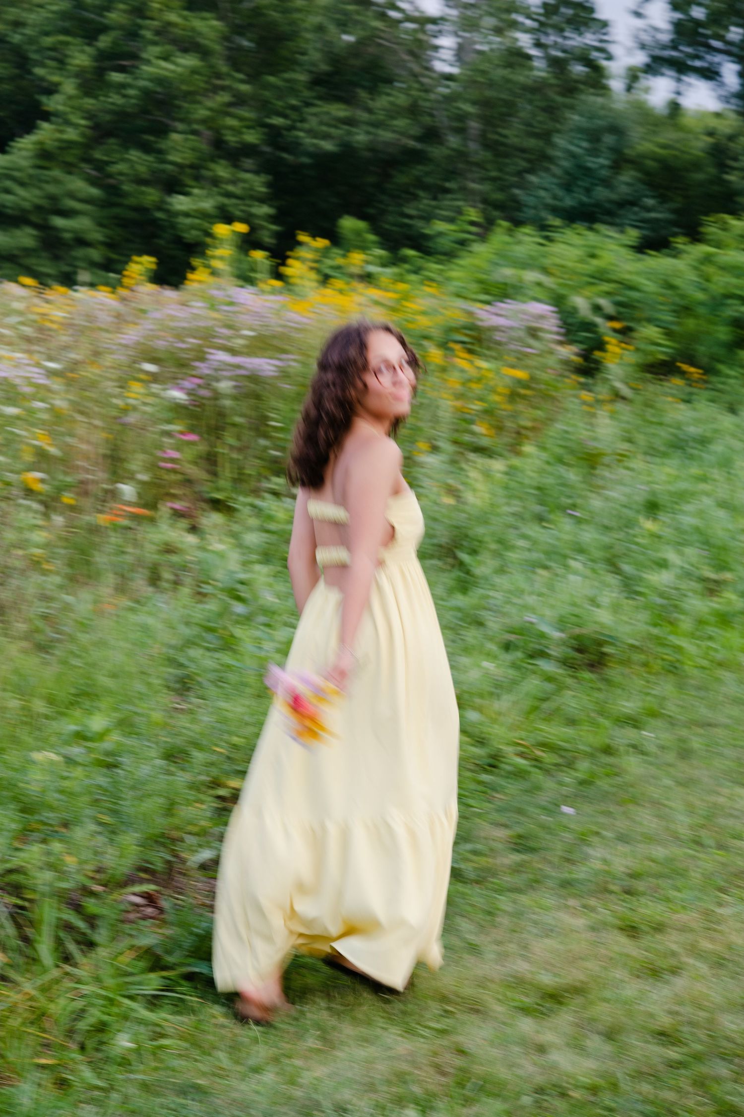 A young woman in a flowing yellow maxi dress walks through a lush green wildflower meadow holding a bouquet.