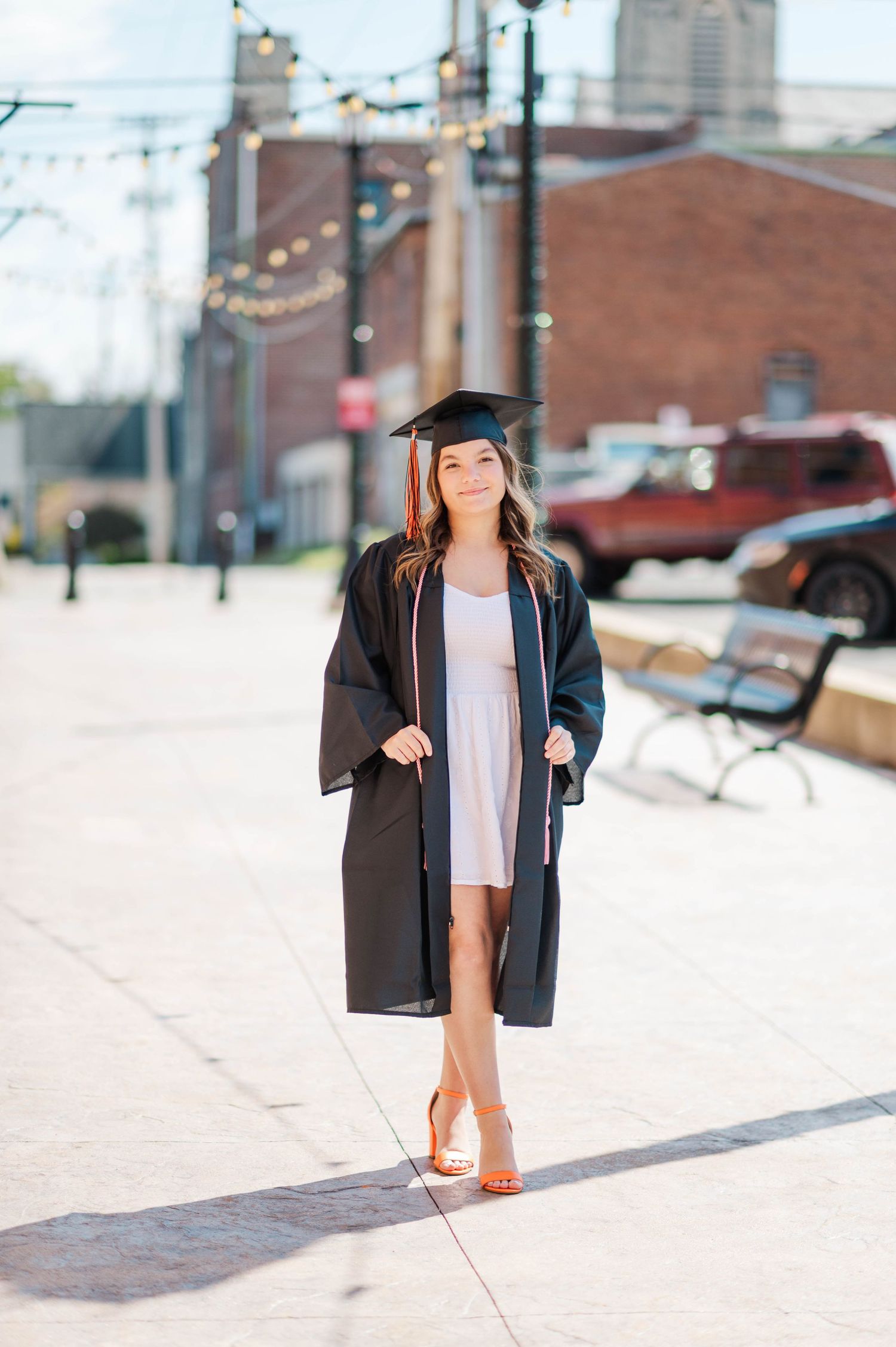 Confident graduate in black cap and gown strolling on sunny urban sidewalk wearing heels.
