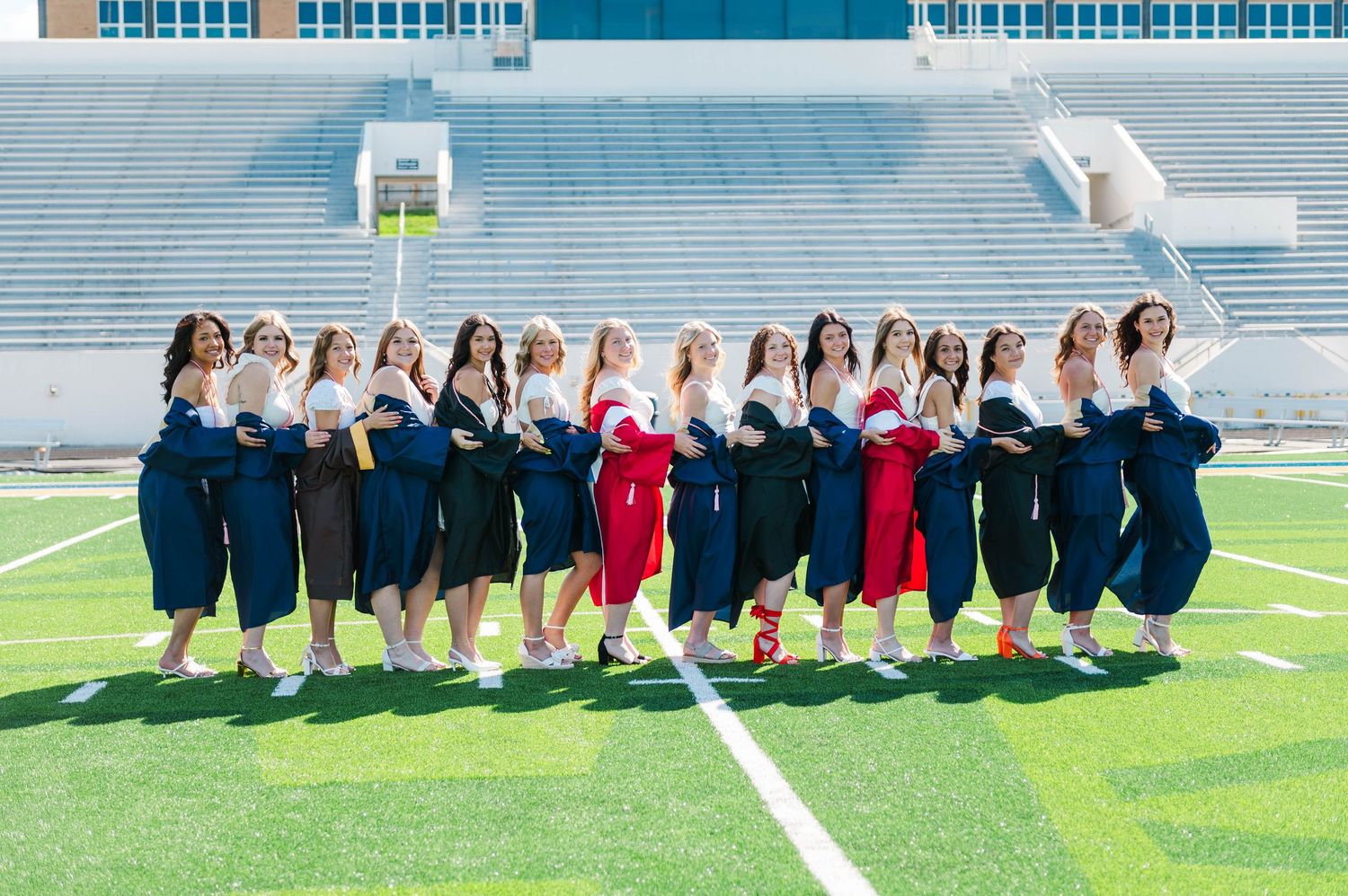 Group of graduates in navy blue gowns tossing caps on a football field, celebrating their graduation day.