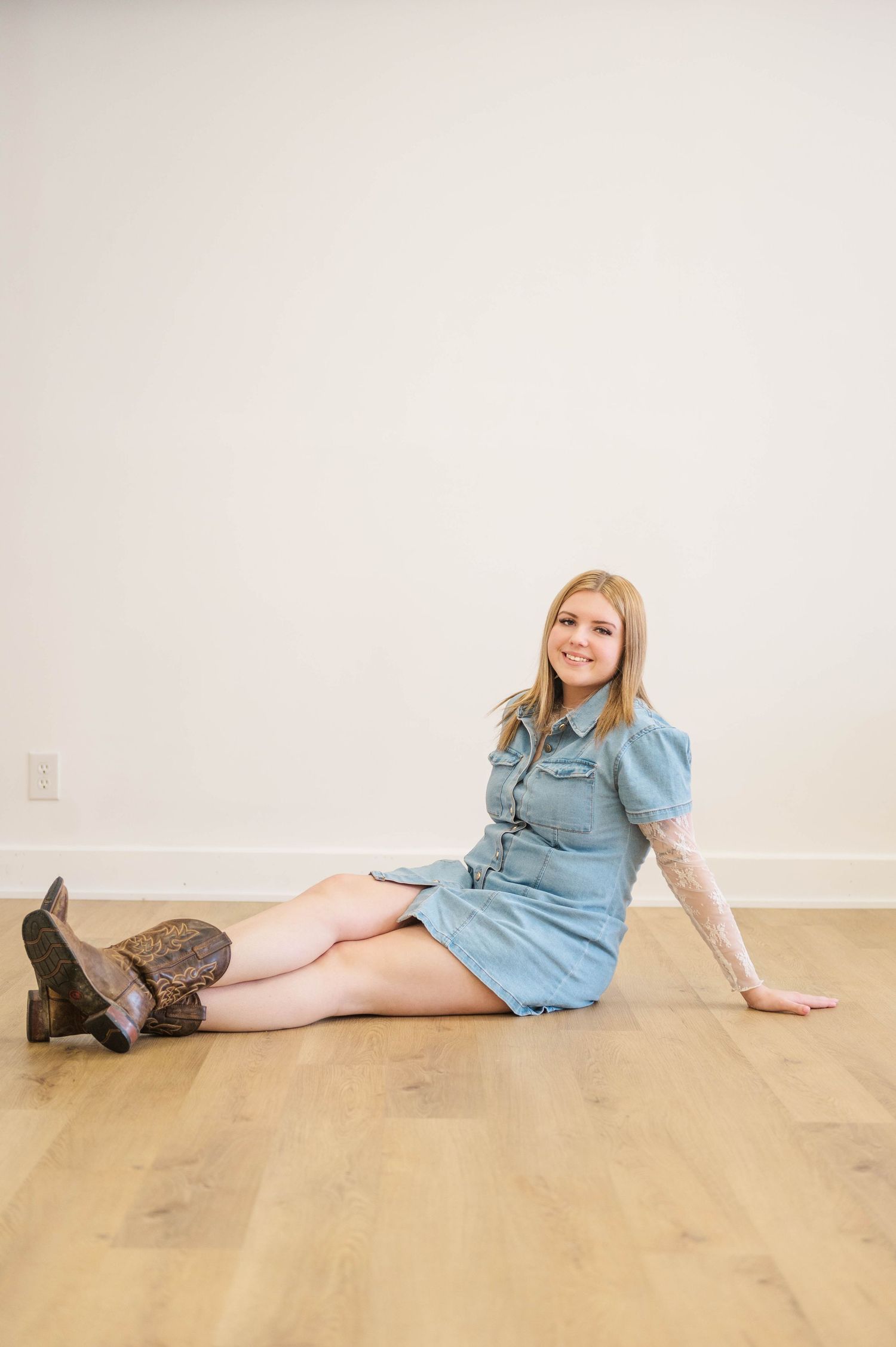 Smiling blonde woman in denim dress and cowboy boots sitting on hardwood floor against white wall.