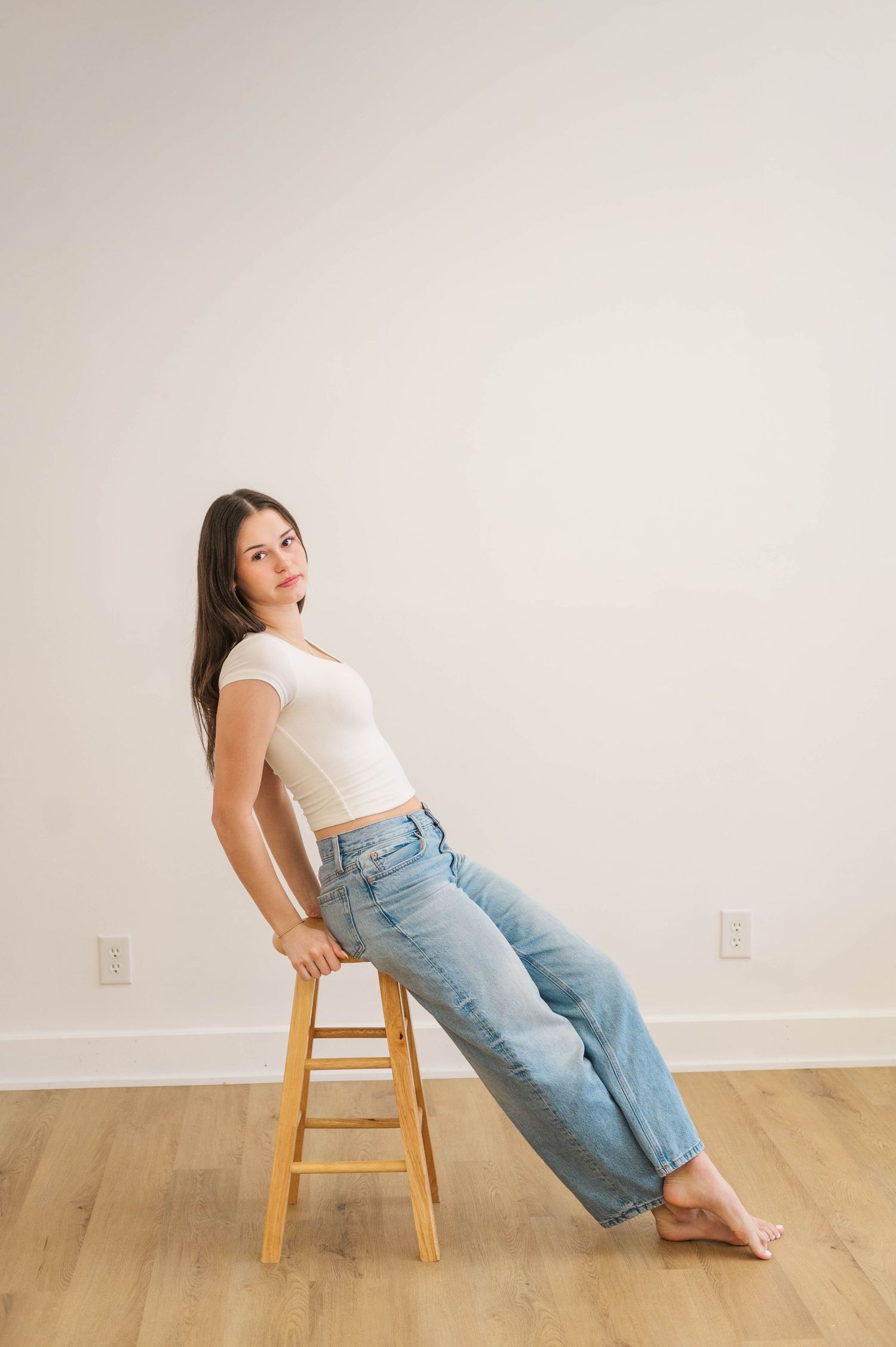 Young woman in white crop top and blue jeans poses barefoot on wooden stool against white wall.