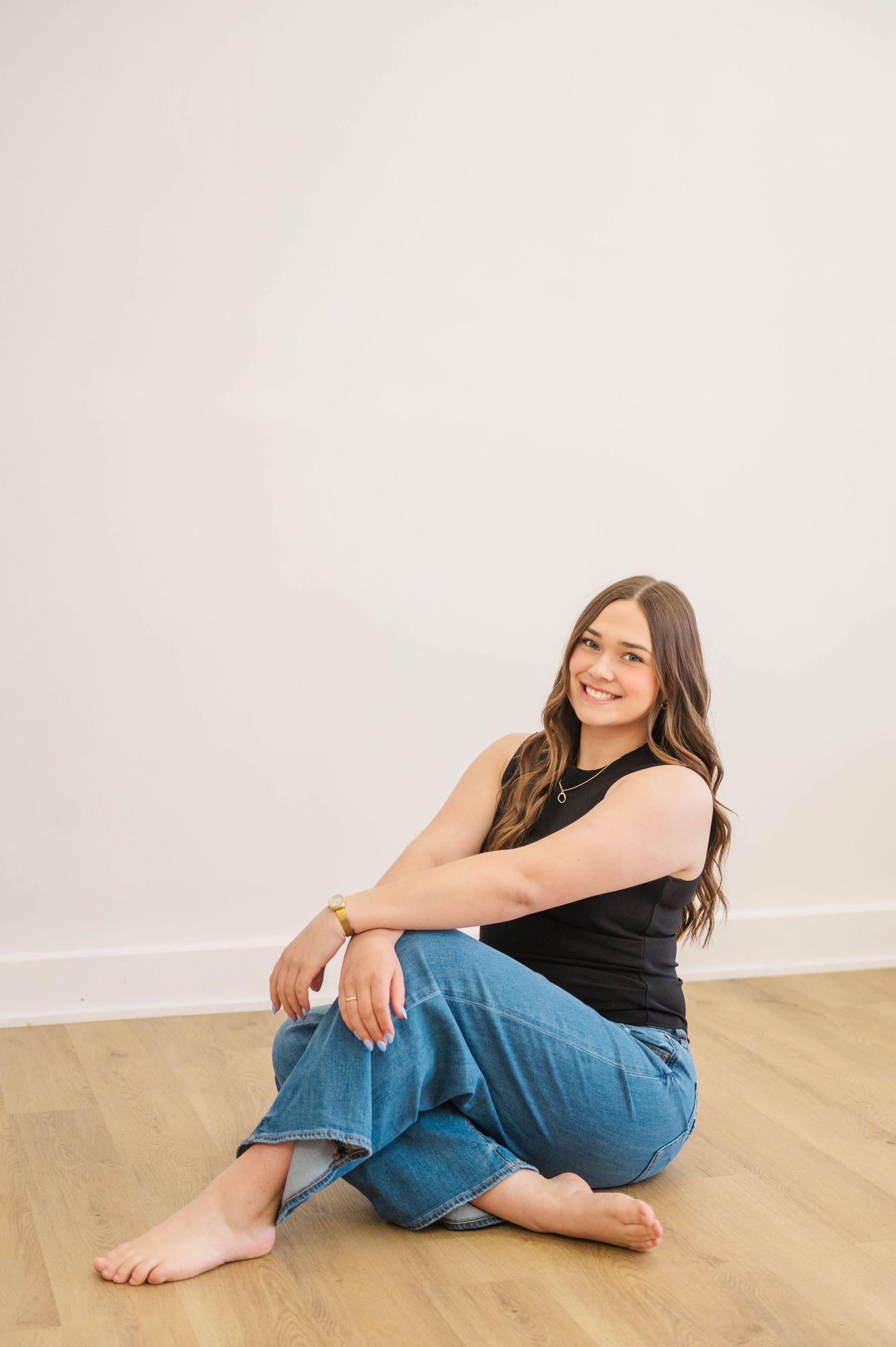Smiling brunette woman sits cross-legged on hardwood floor wearing black sleeveless top and wide-leg blue jeans.