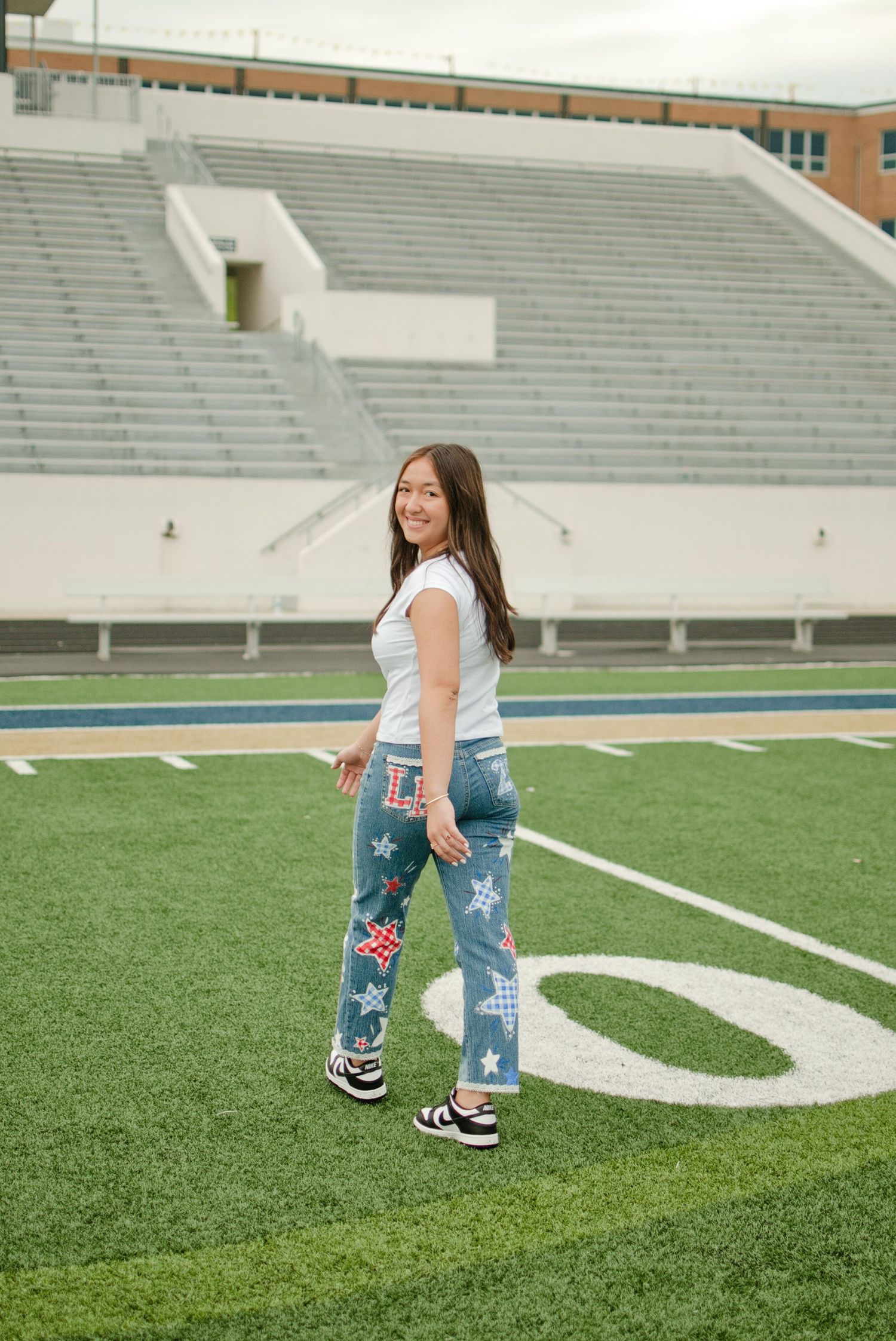 Teen girl in custom star-painted jeans and white tee smiles sideways on a football field turf.