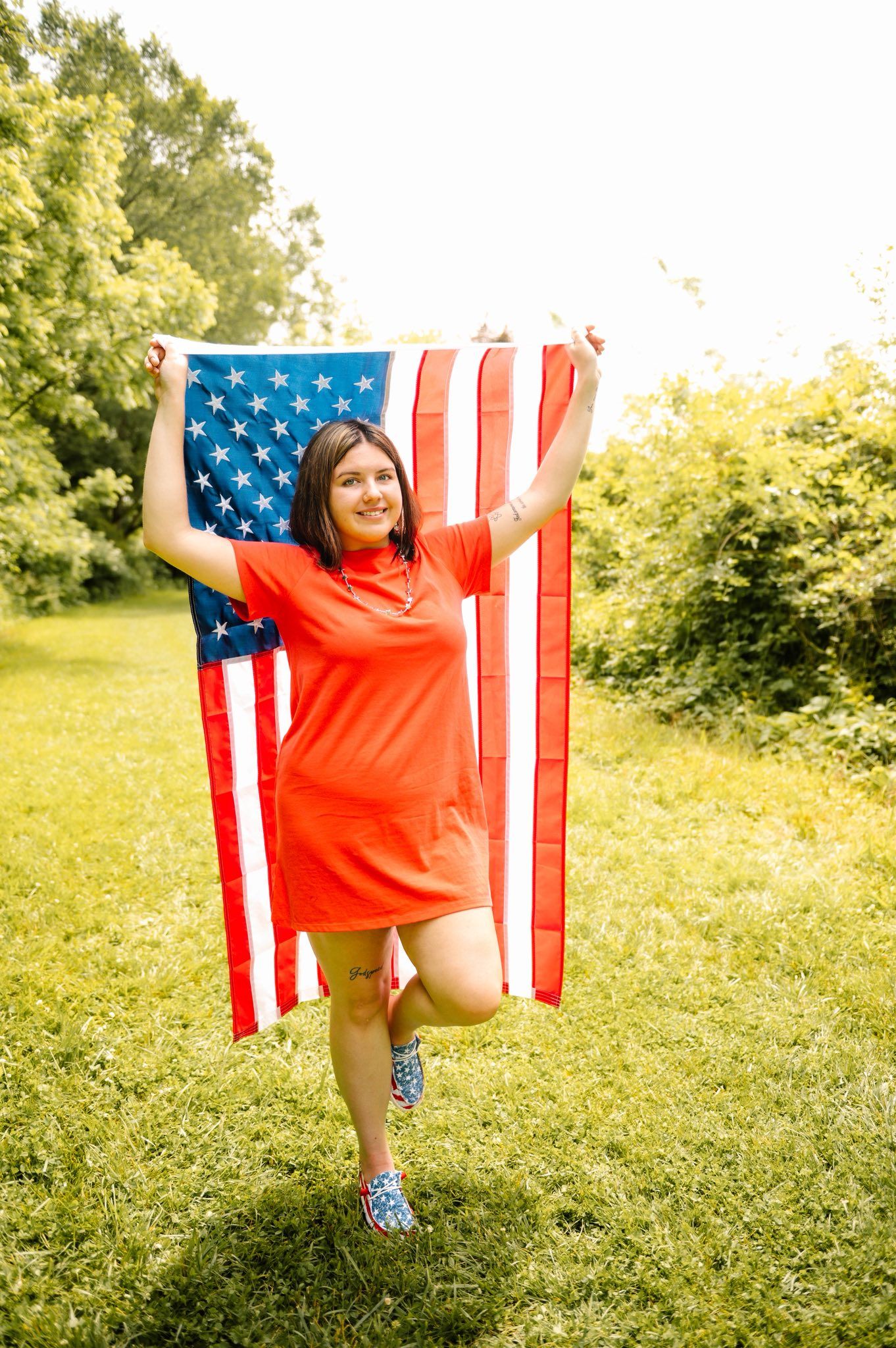 Person in red shirt poses with American flag against green summer backdrop.