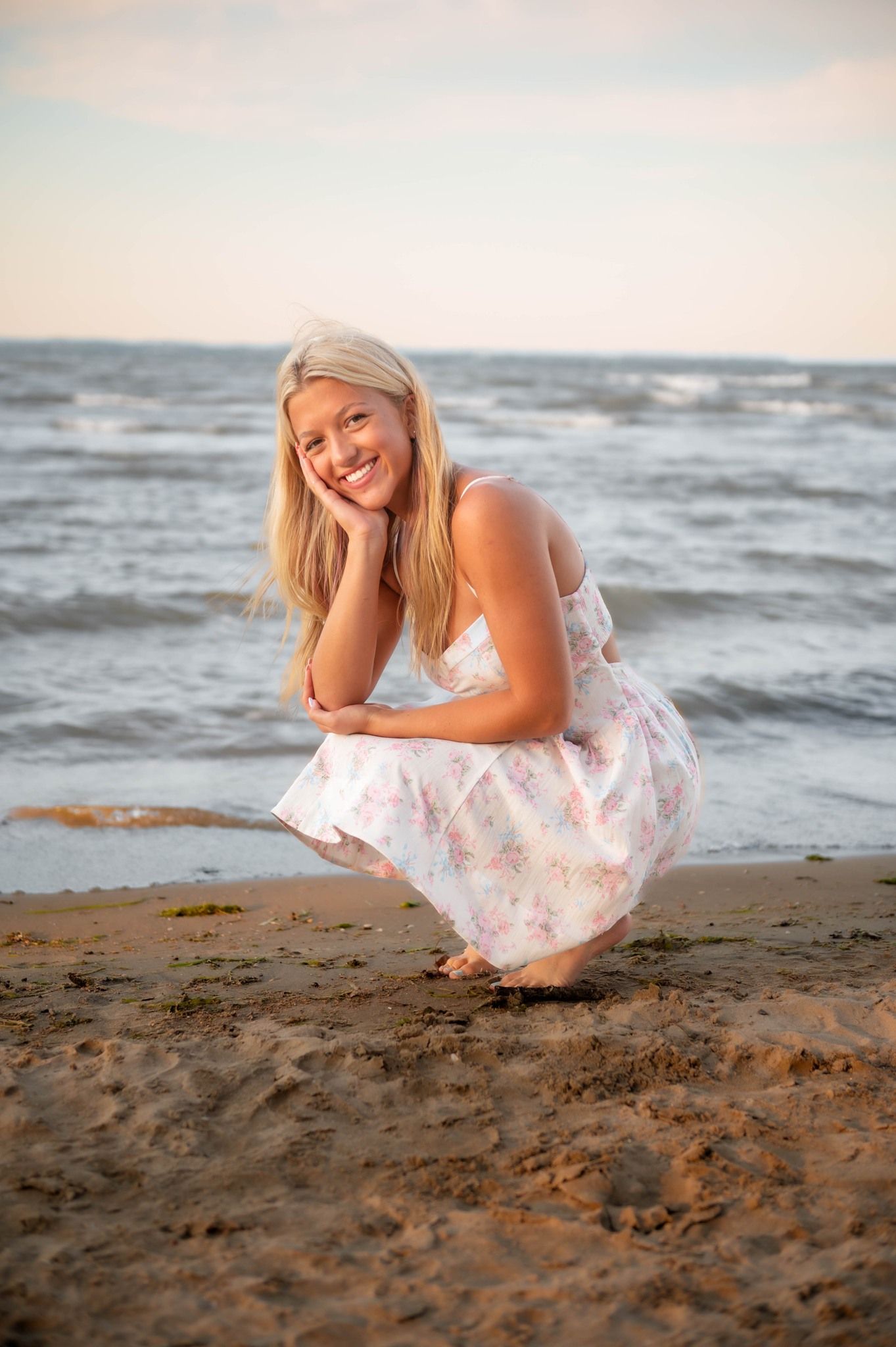 Woman in floral dress crouching on sandy beach at sunset.