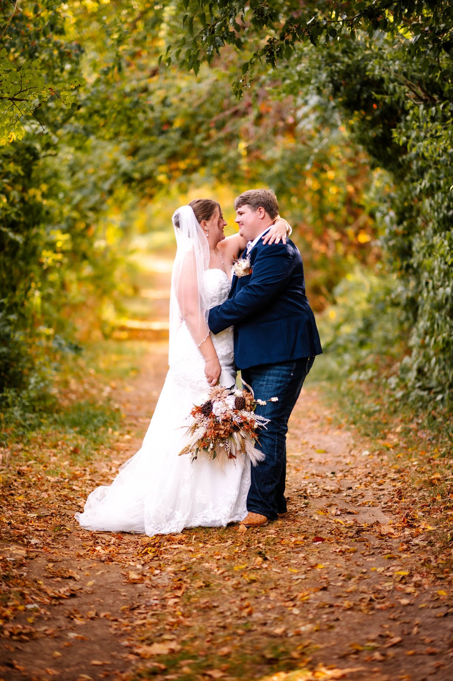 Couple shares romantic moment on leaf-covered autumn path surrounded by trees.