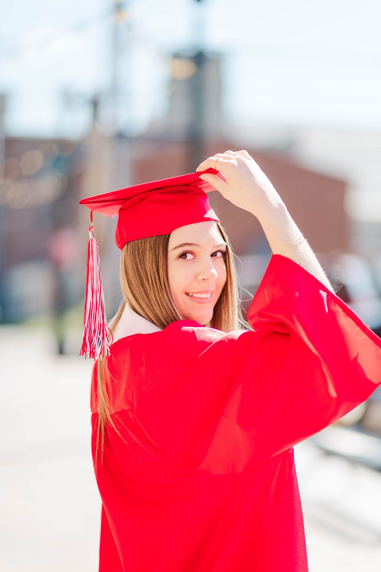 Young woman in red graduation cap and gown smiling over shoulder while adjusting cap outdoors.