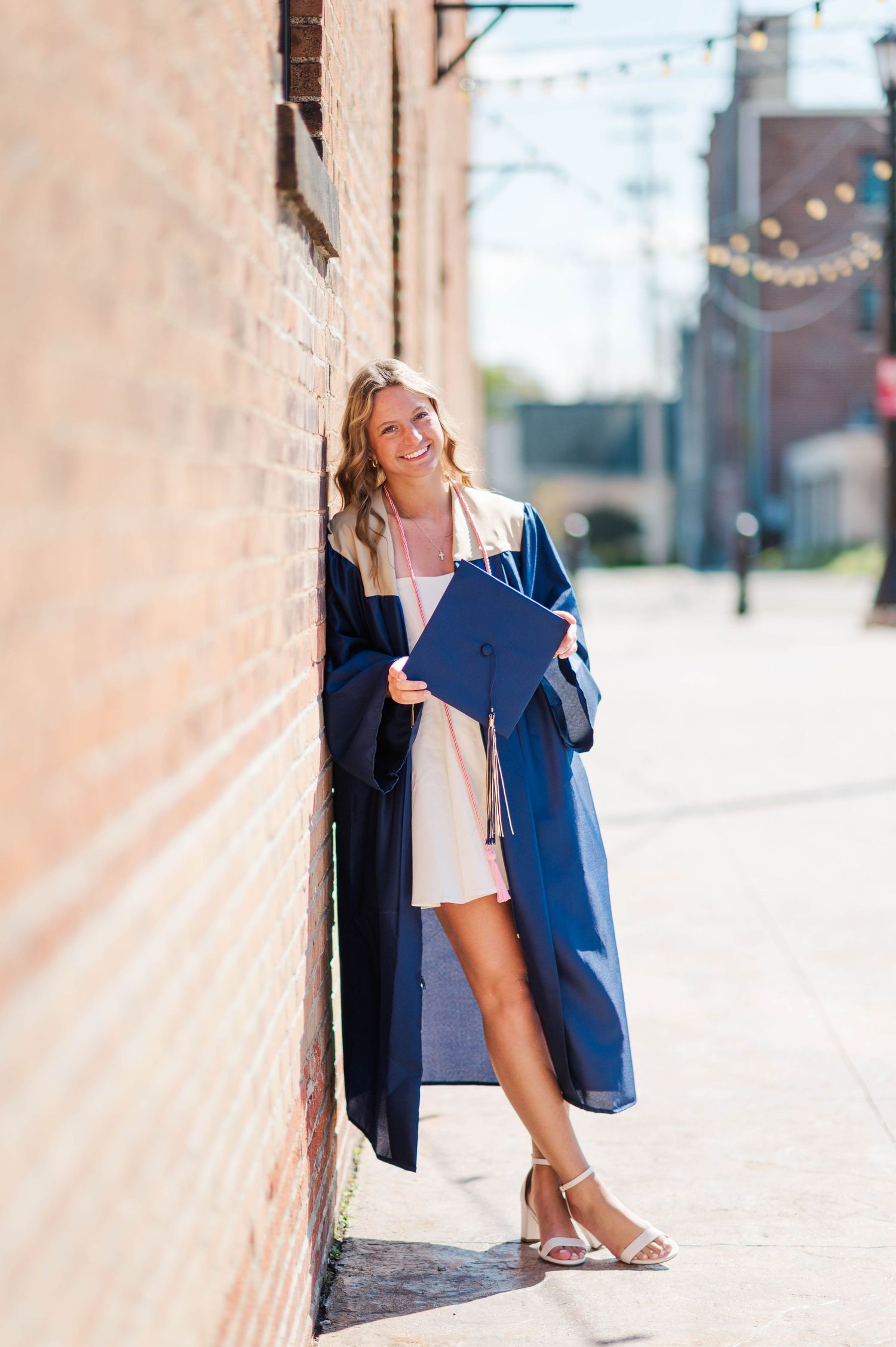 Smiling graduate in navy blue cap and gown leaning against brick wall holding diploma outdoors.