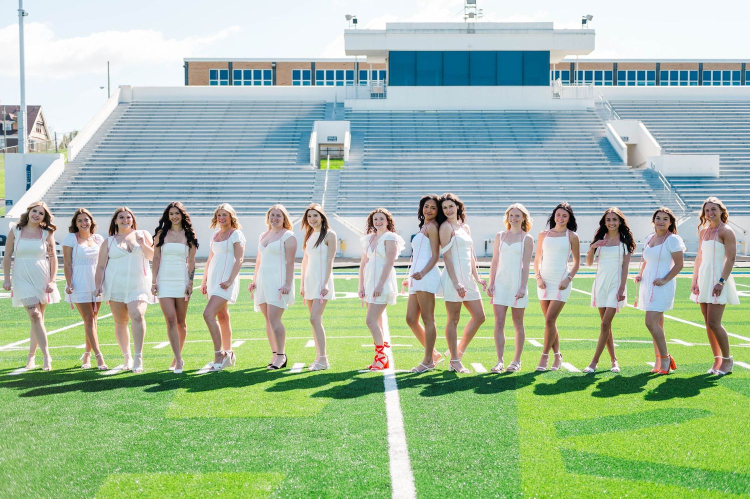 Group of young women in white dresses posing on a football field with stadium bleachers in the background.