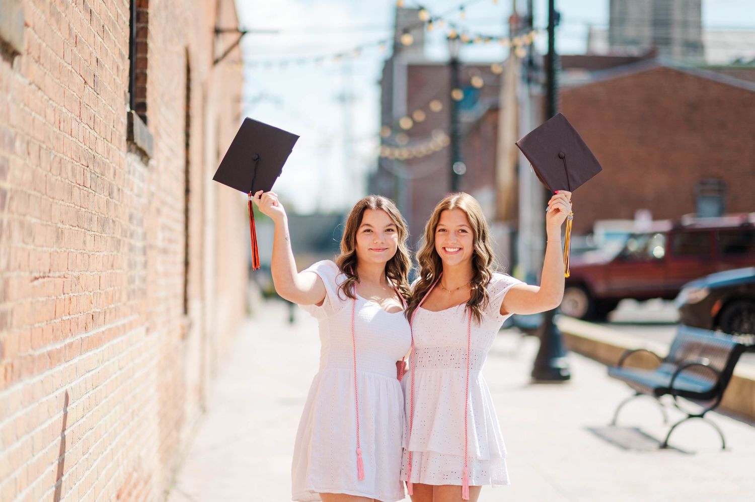 Two smiling female graduates in white dresses raising graduation caps outdoors on sunny day.