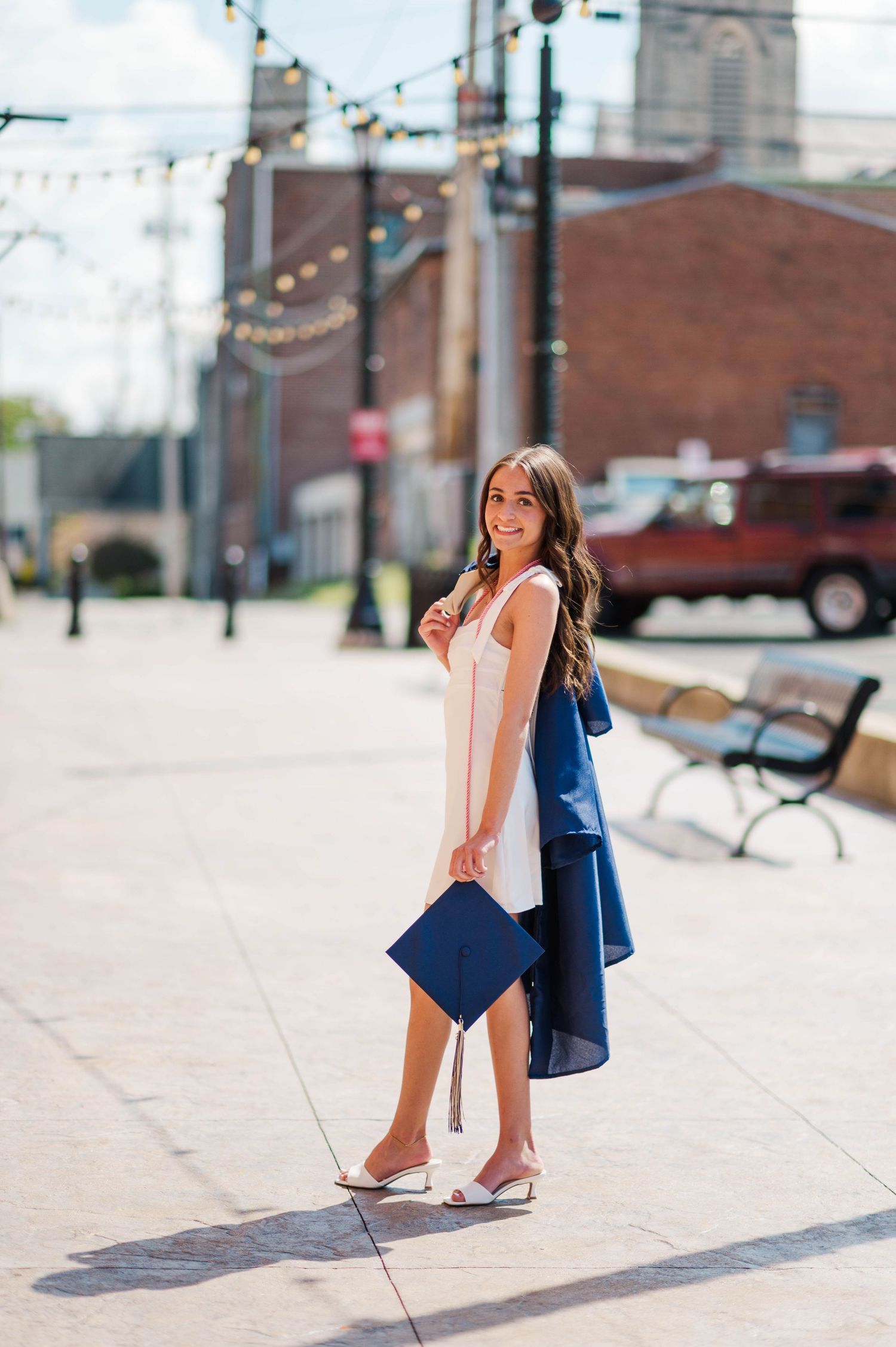 Smiling brunette woman in white dress carrying navy graduation cap and gown on sunny street.