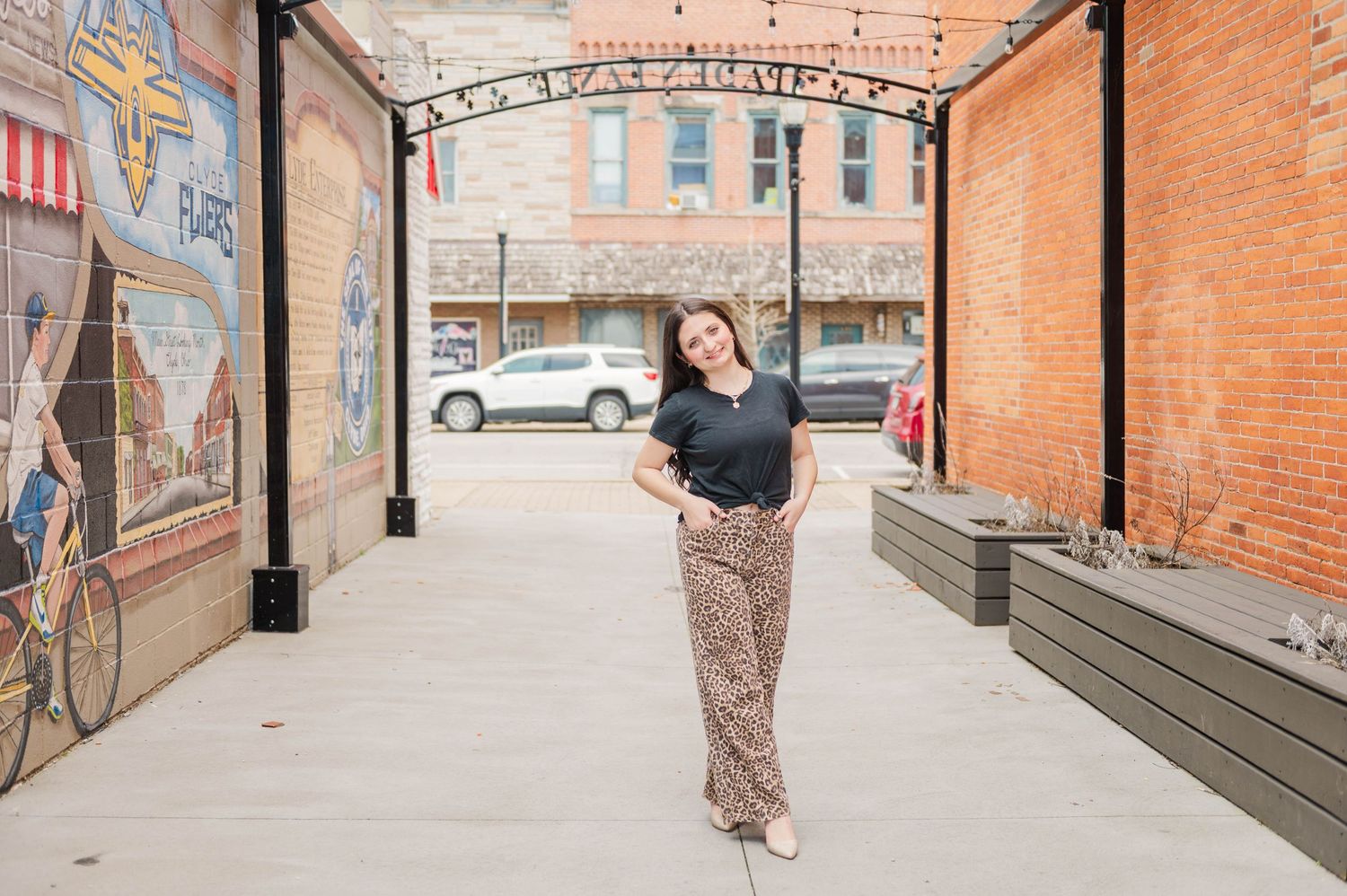 Woman in black top and leopard print pants smiling while posing in an urban alley with a colorful mural.