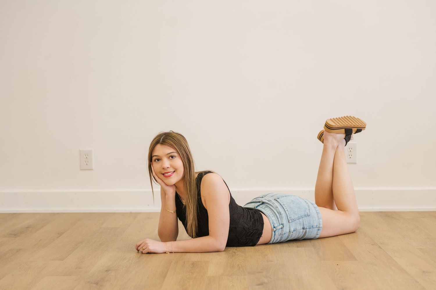 Teen girl in black tank and denim shorts lying on hardwood floor, smiling at camera.