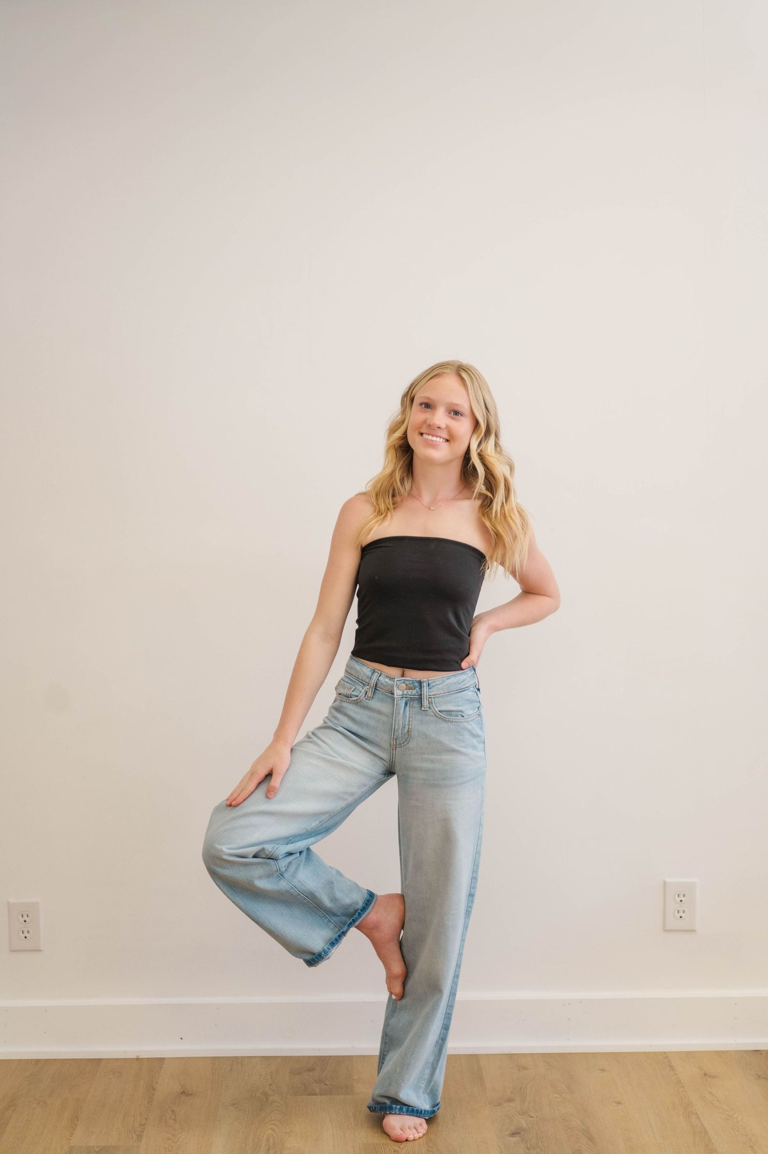 Smiling blonde woman in black tube top and wide-leg jeans posing barefoot on one leg against white wall.