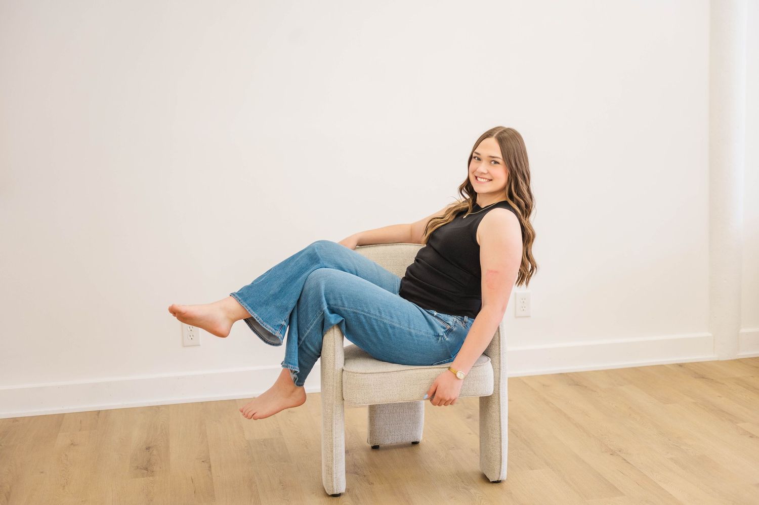 Woman in black top and jeans sitting relaxed in white chair, smiling, on hardwood floor.