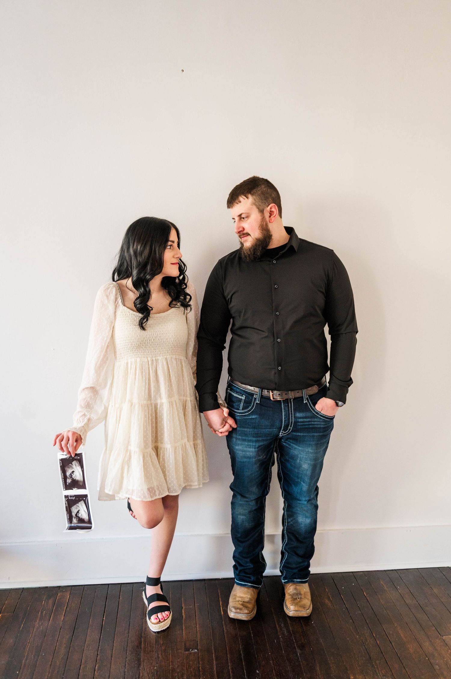 A couple in casual attire stands together on hardwood floors holding pregnancy announcement photos.