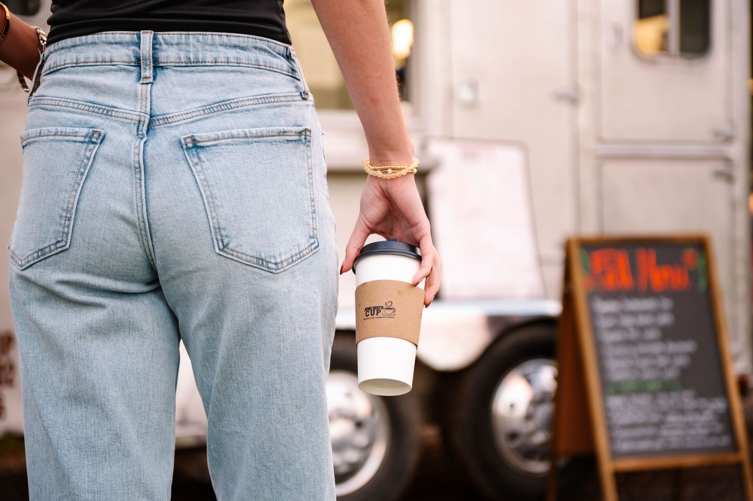 Person wearing light blue jeans and black top holding coffee cup near food truck.