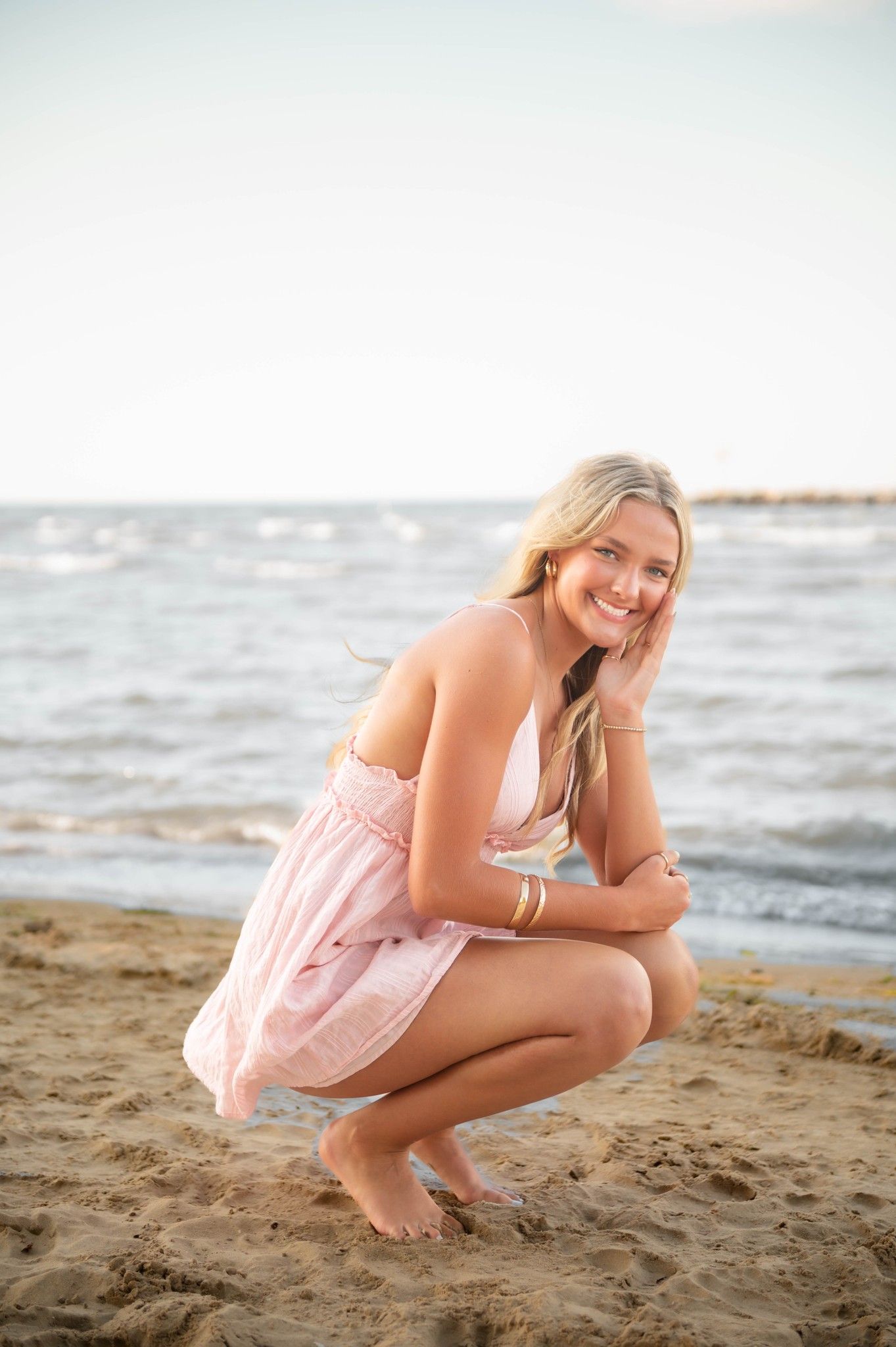 Woman in pink dress crouching on sandy beach with ocean waves in background.