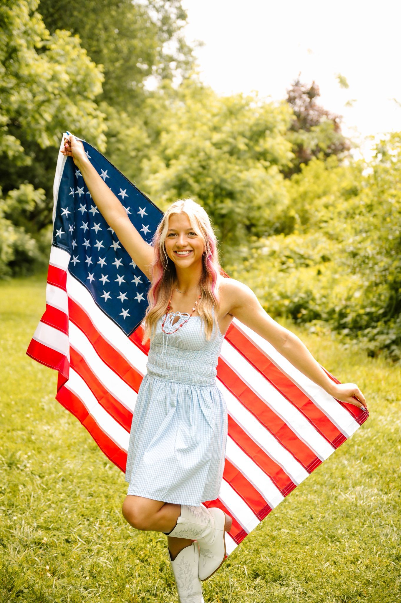 Person in light dress jumps joyfully while holding American flags in sunny field.