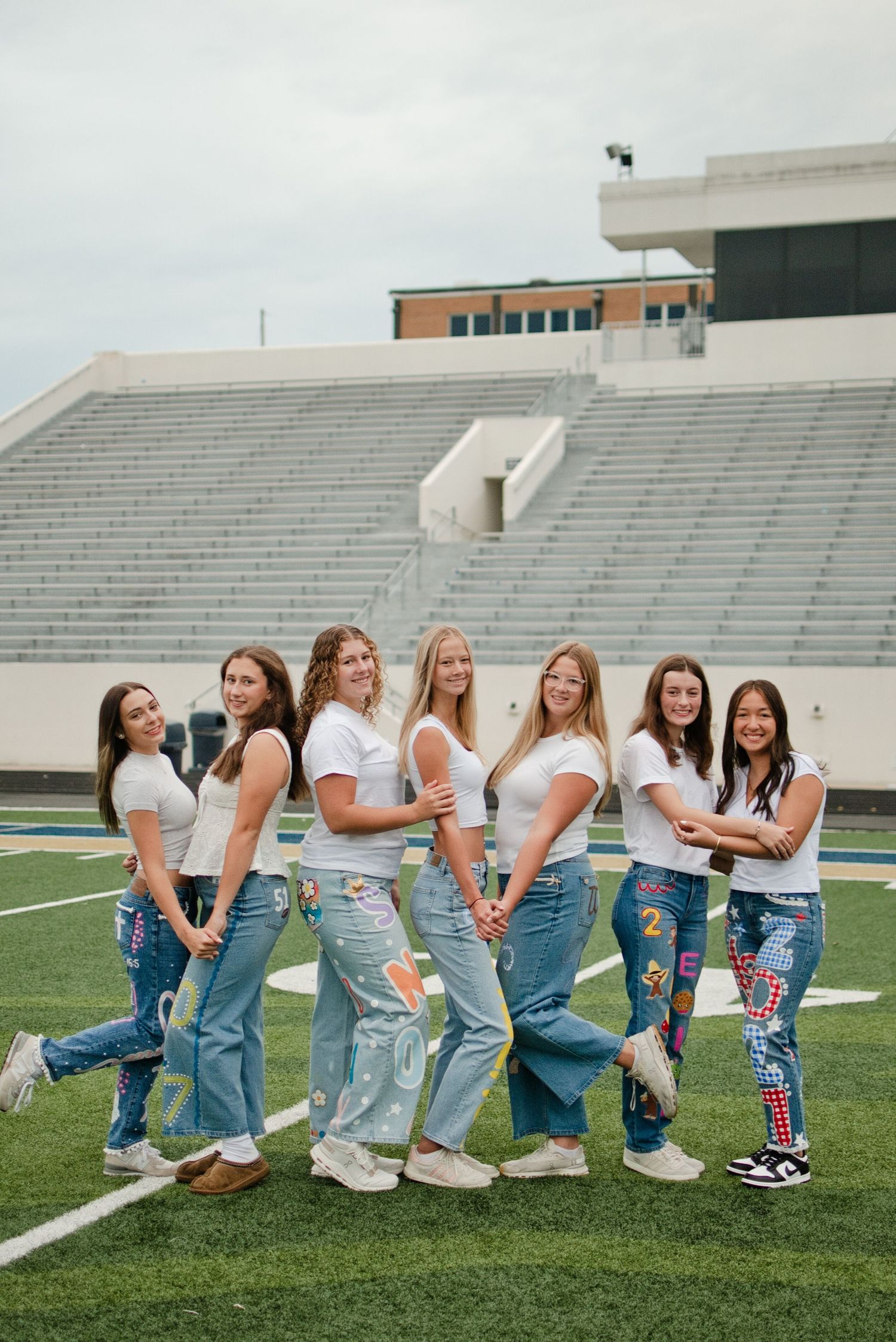 Seven young women pose together on a football field wearing white tops and decorated denim jeans.