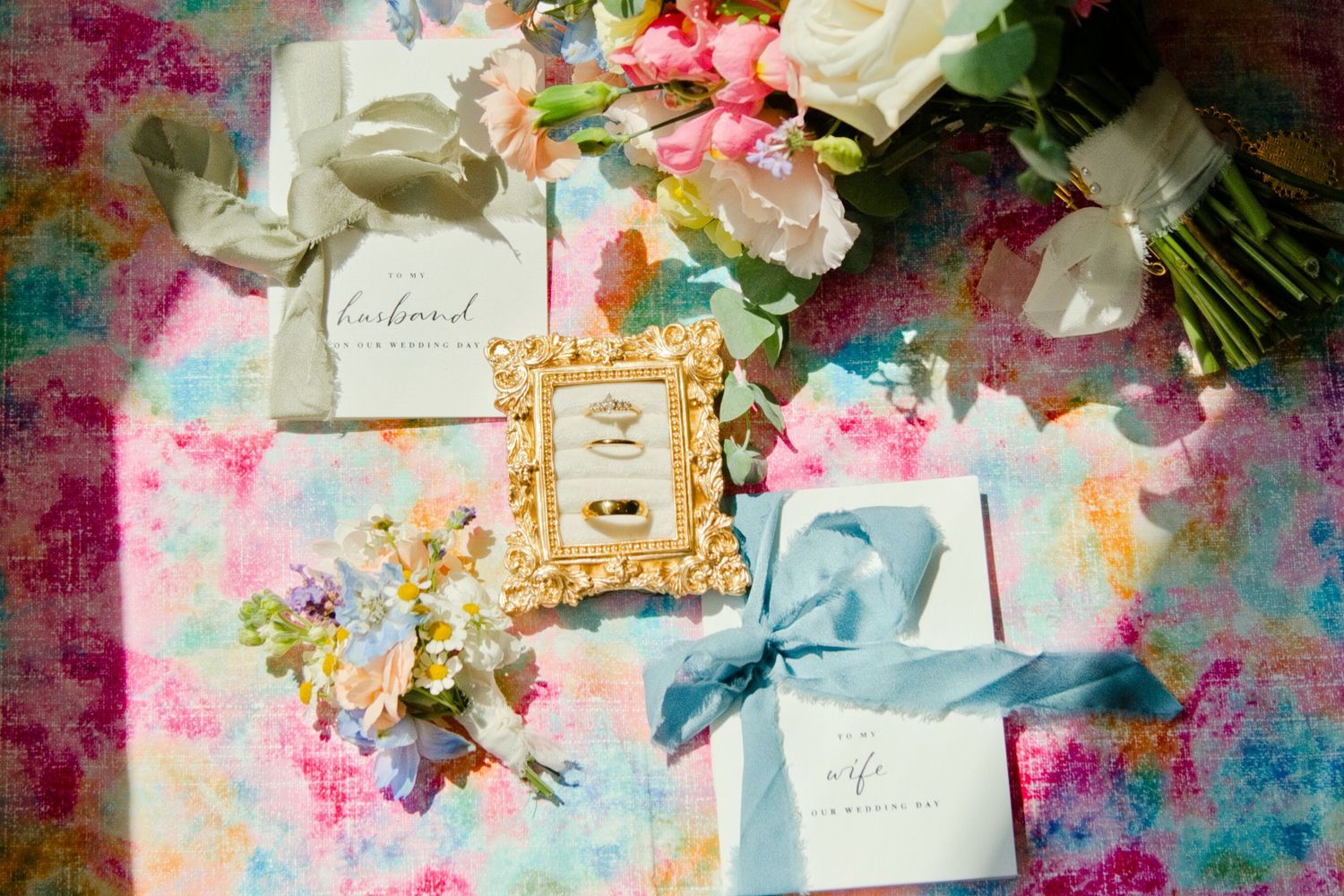 Elegant wedding flat lay with gold frame, rings, blue ribbon gift box, floral bouquet, and colorful pastel background.