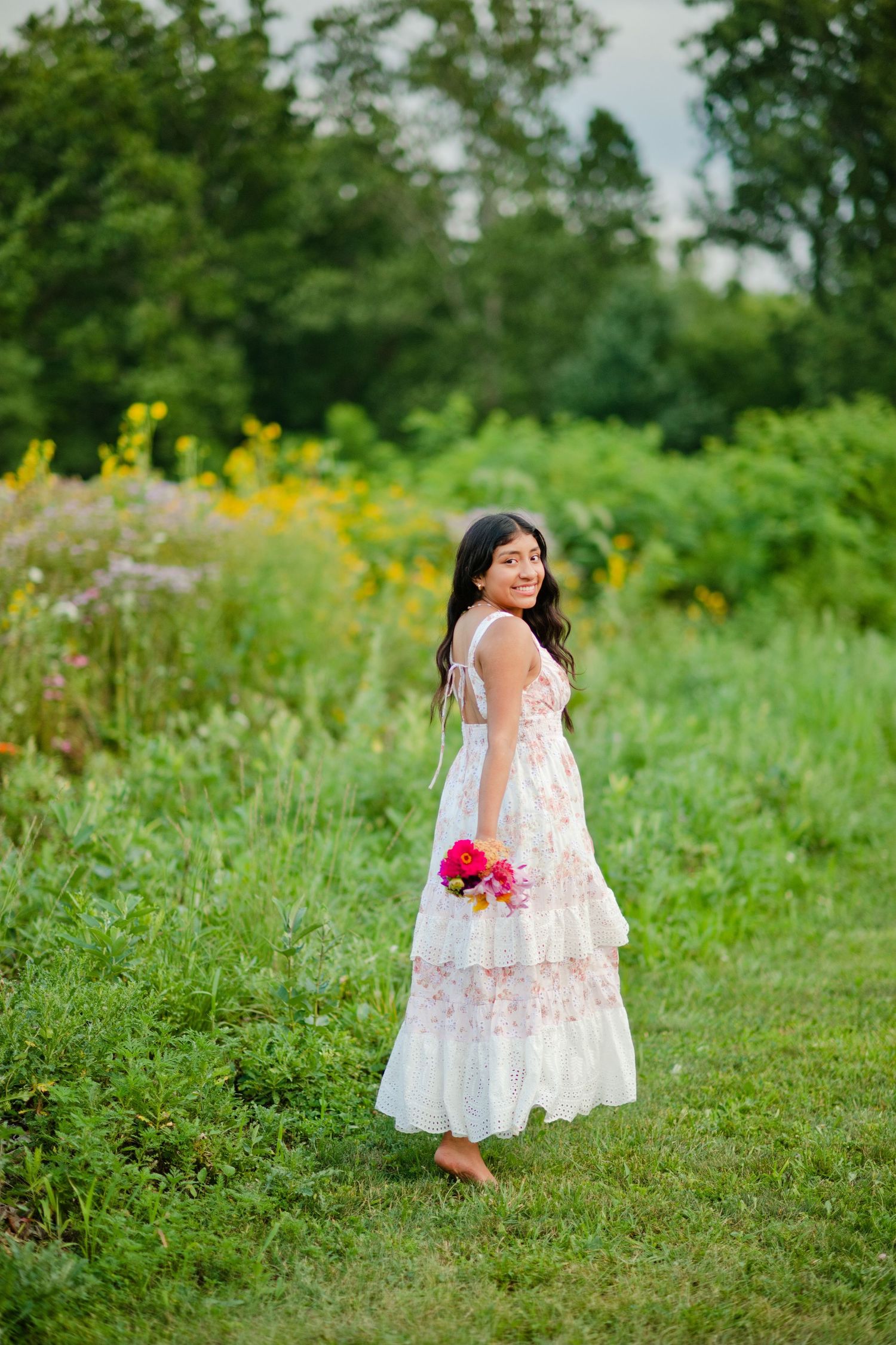 Young woman in white floral dress holding pink flowers, smiling in a lush green meadow.
