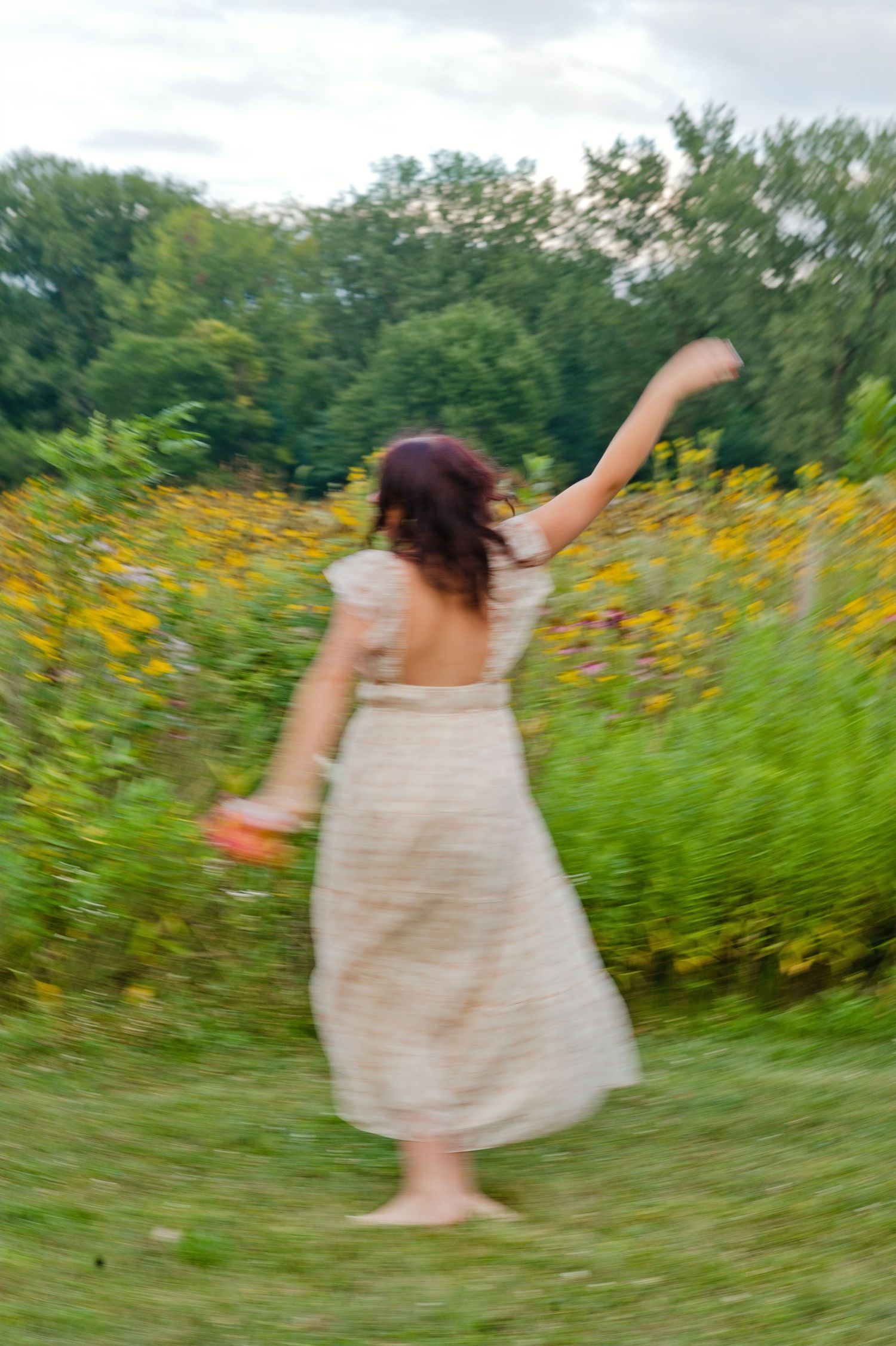 A woman in a white backless dress twirls barefoot in a lush garden filled with yellow wildflowers on a cloudy day.