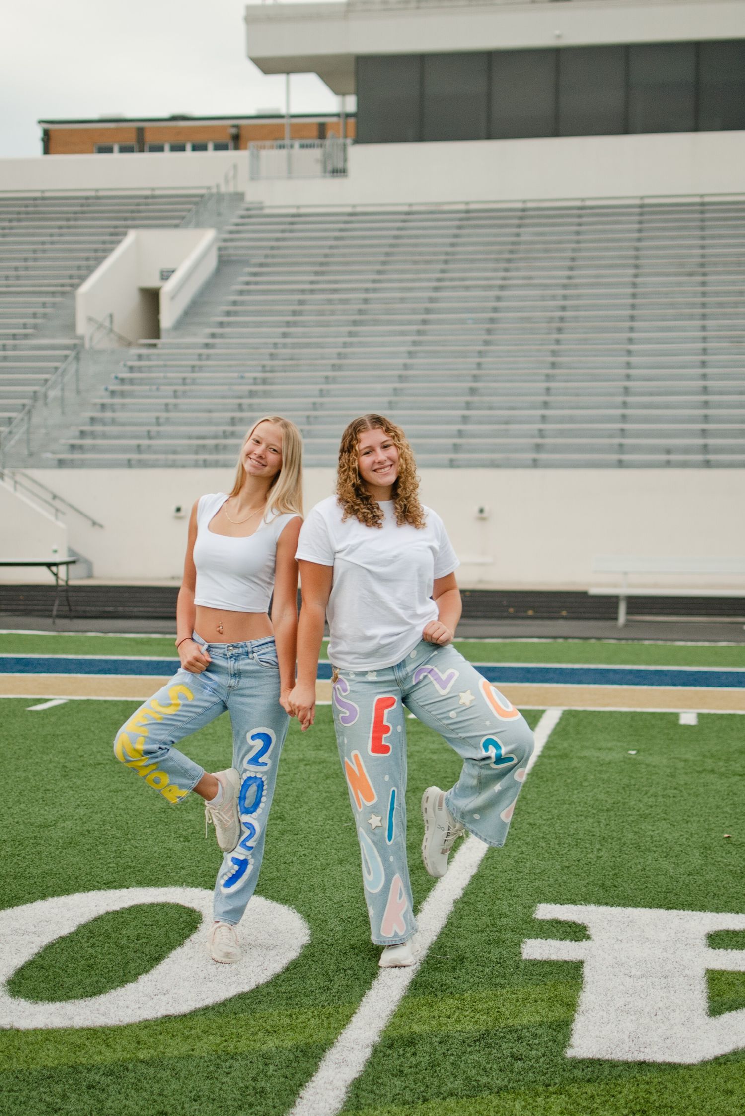 Two smiling girls in white tops and painted senior 2027 jeans pose back-to-back on a football field.