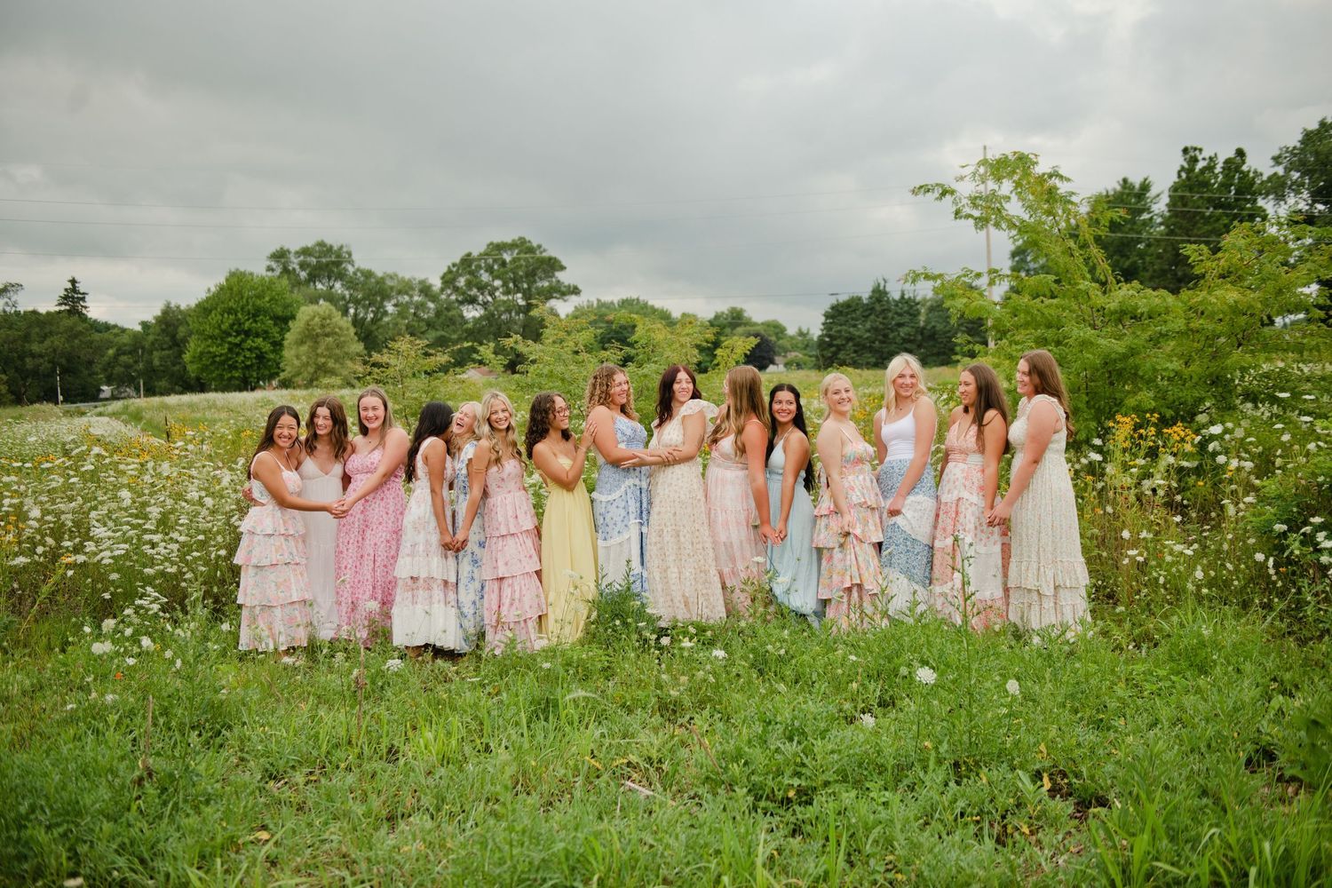 Group of women in pastel maxi dresses posing together in a wildflower meadow under cloudy skies.