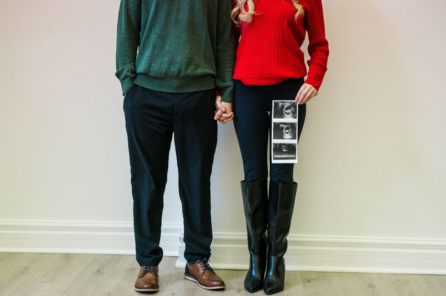 Couple standing together holding ultrasound photos against white wall.