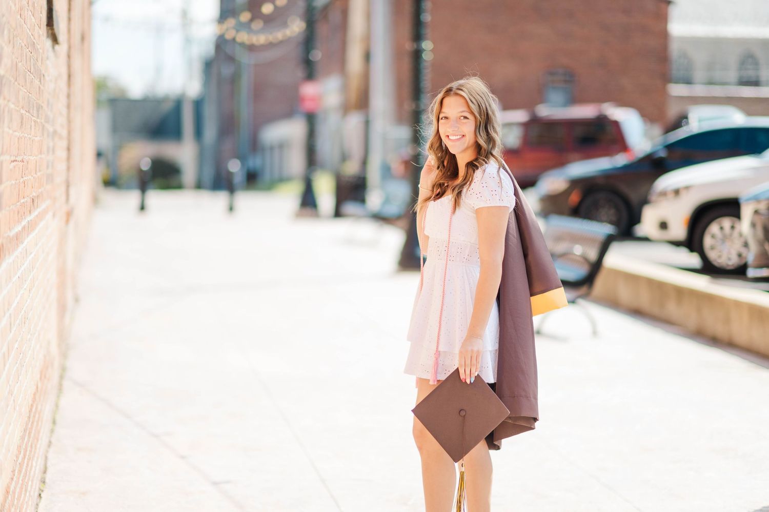 Young woman in pink dress carrying brown graduation gown and cap on sunny street.
