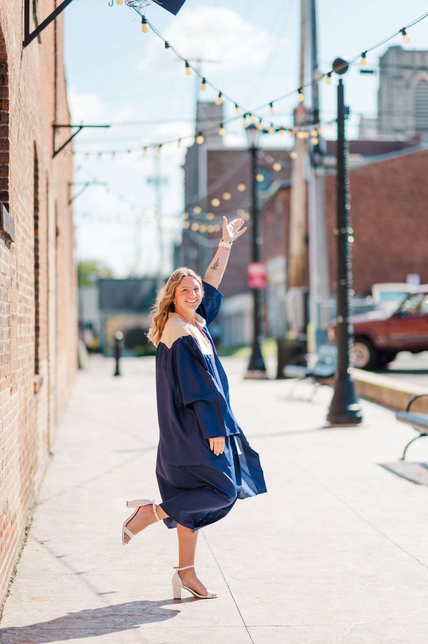 Joyful graduate in navy blue gown tosses her cap in the air on a charming city sidewalk.