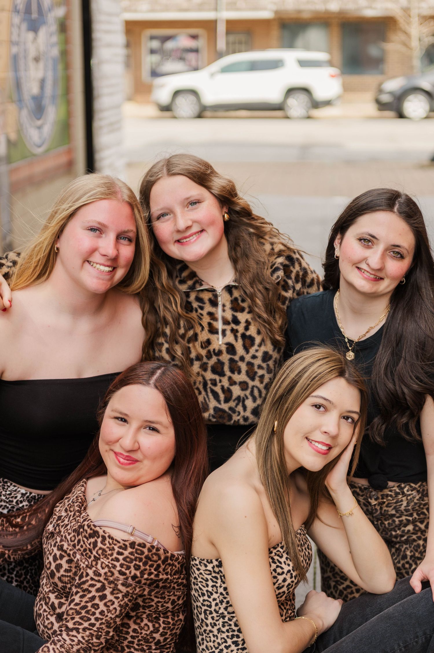 Five young women smiling together outdoors, wearing leopard print and black outfits in urban setting.