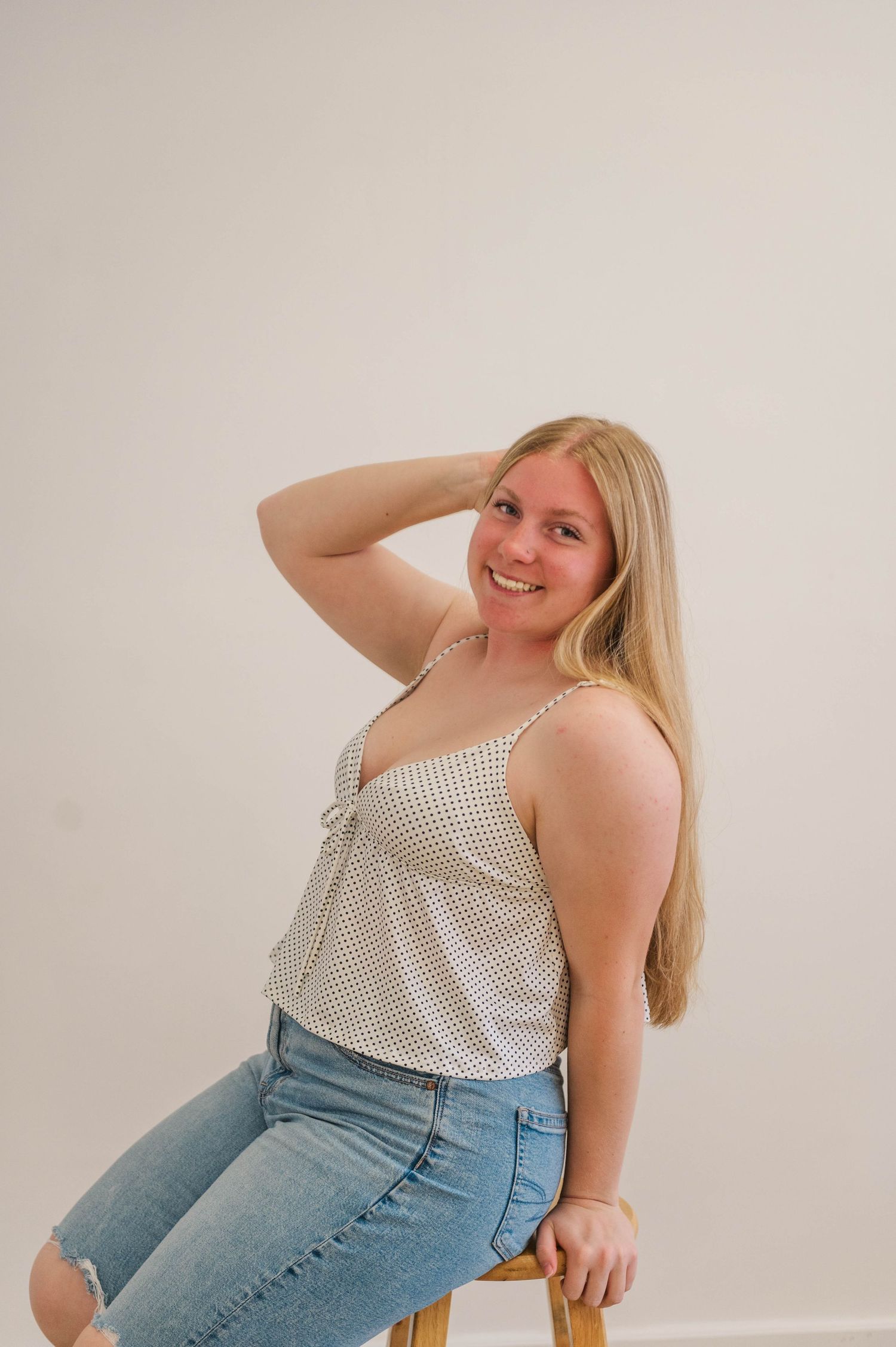 Smiling blonde woman in polka dot tank top and ripped jeans sits on stool with hand in hair.