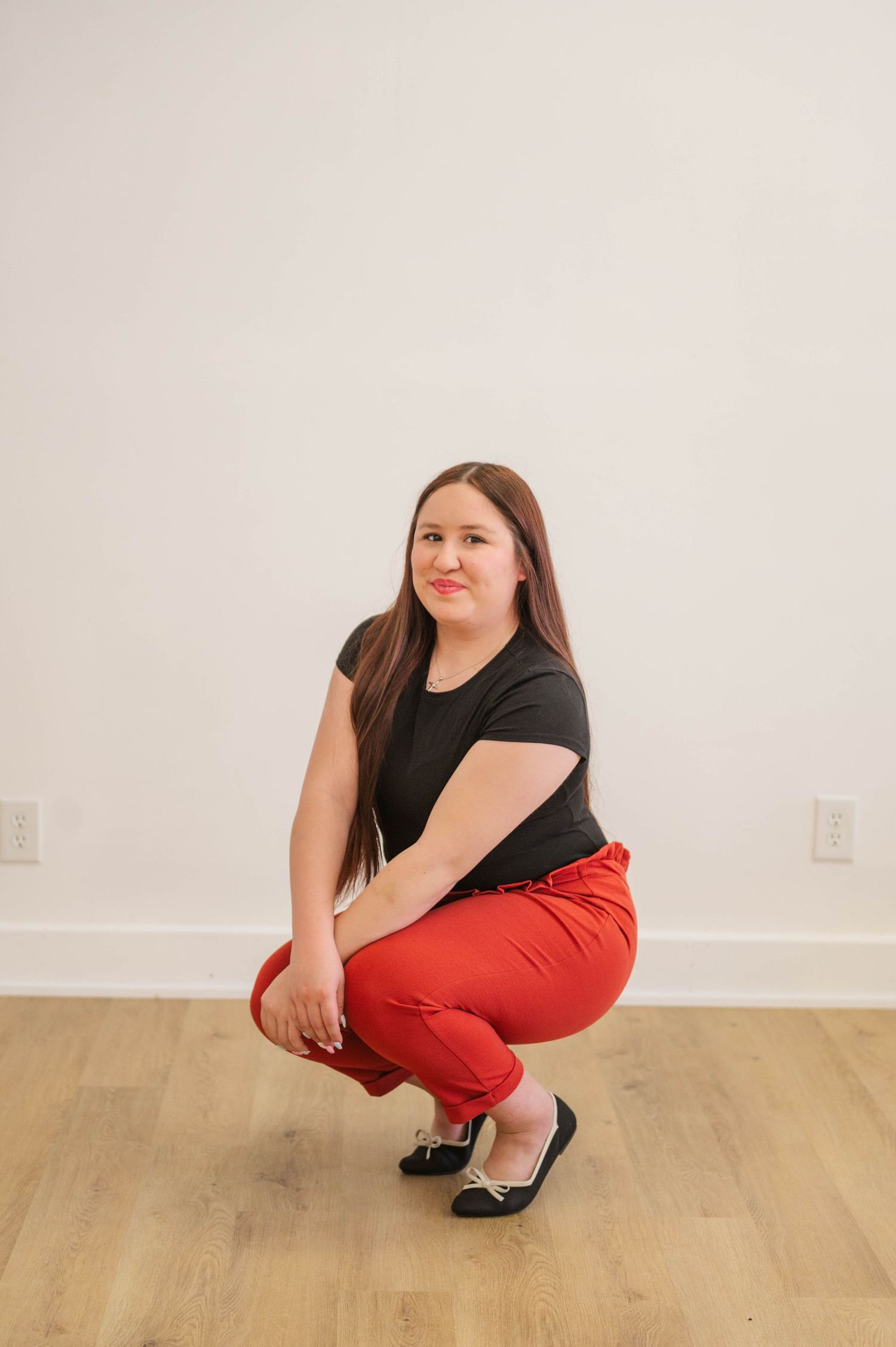 Young woman in black top and red pants crouching on wood floor against white wall.