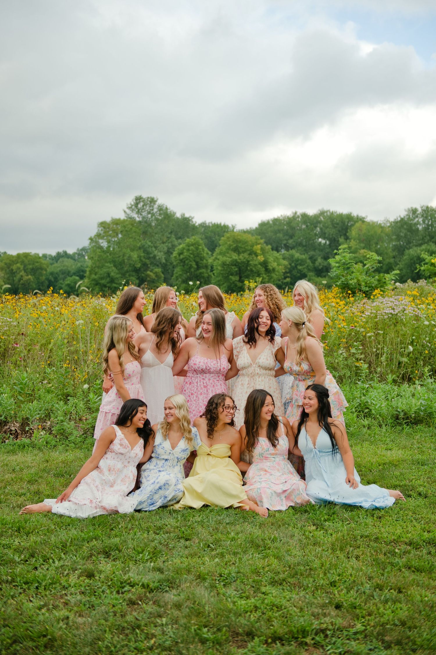 Group of twelve women in pastel floral dresses laughing together in a wildflower meadow.