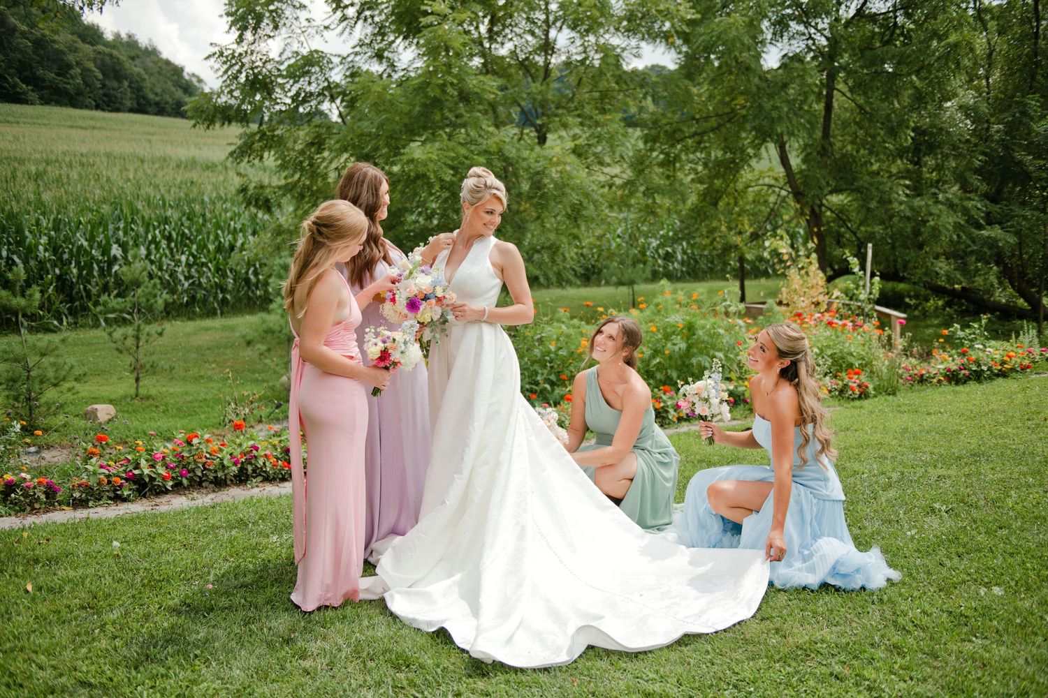 Bride in white gown with bridesmaids in pink and blue dresses posing in a lush garden setting.