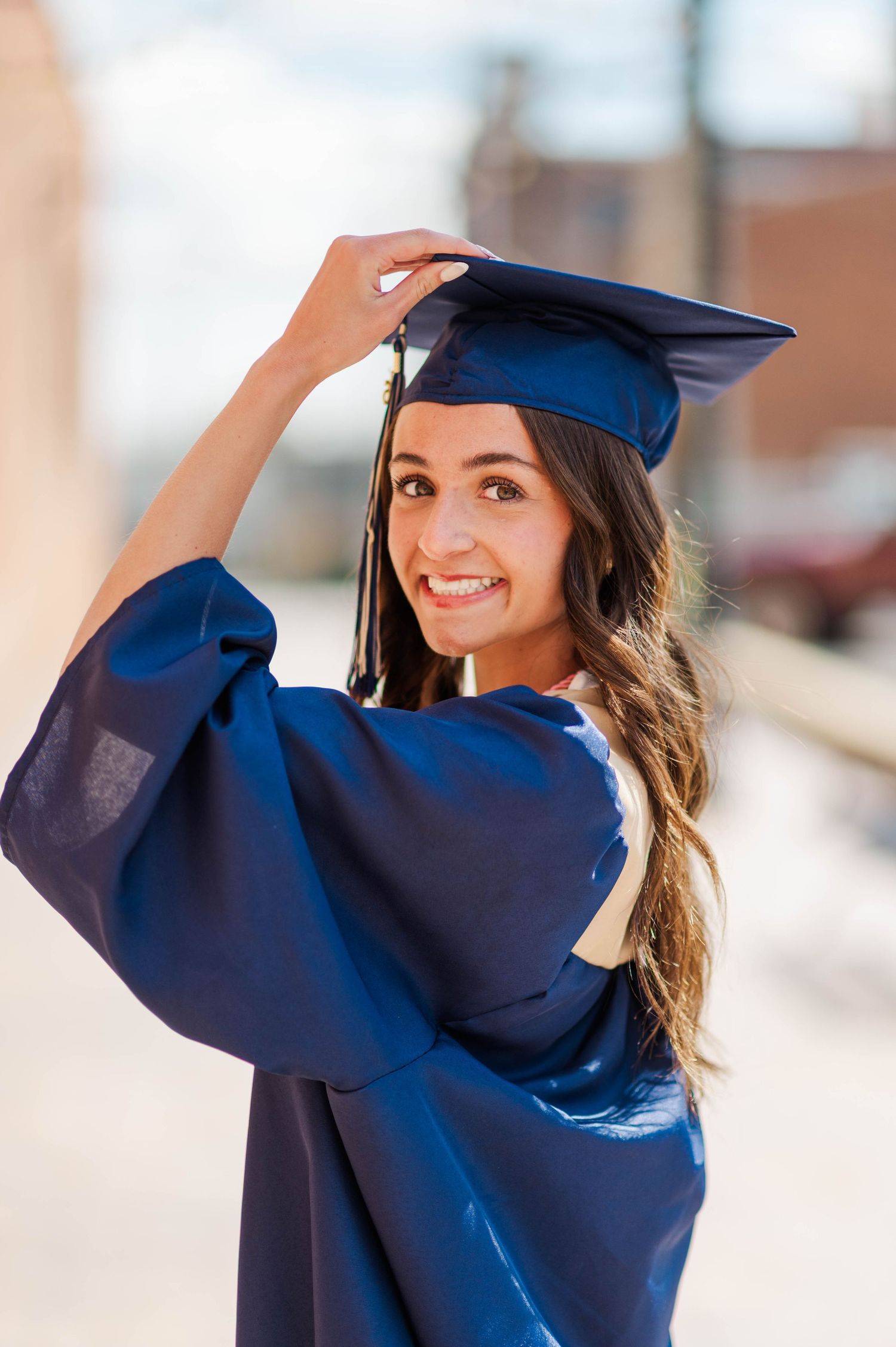 Smiling graduate in navy blue cap and gown adjusting mortarboard while posing outdoors.