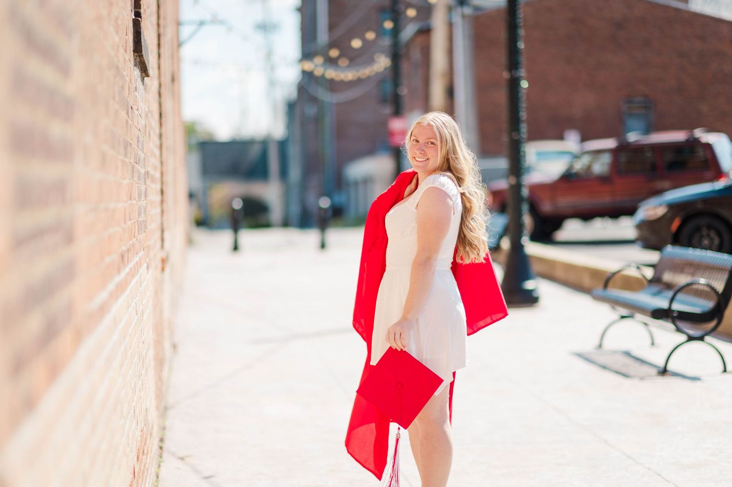 Blonde woman in white dress with red graduation gown on sunny brick-lined city sidewalk.