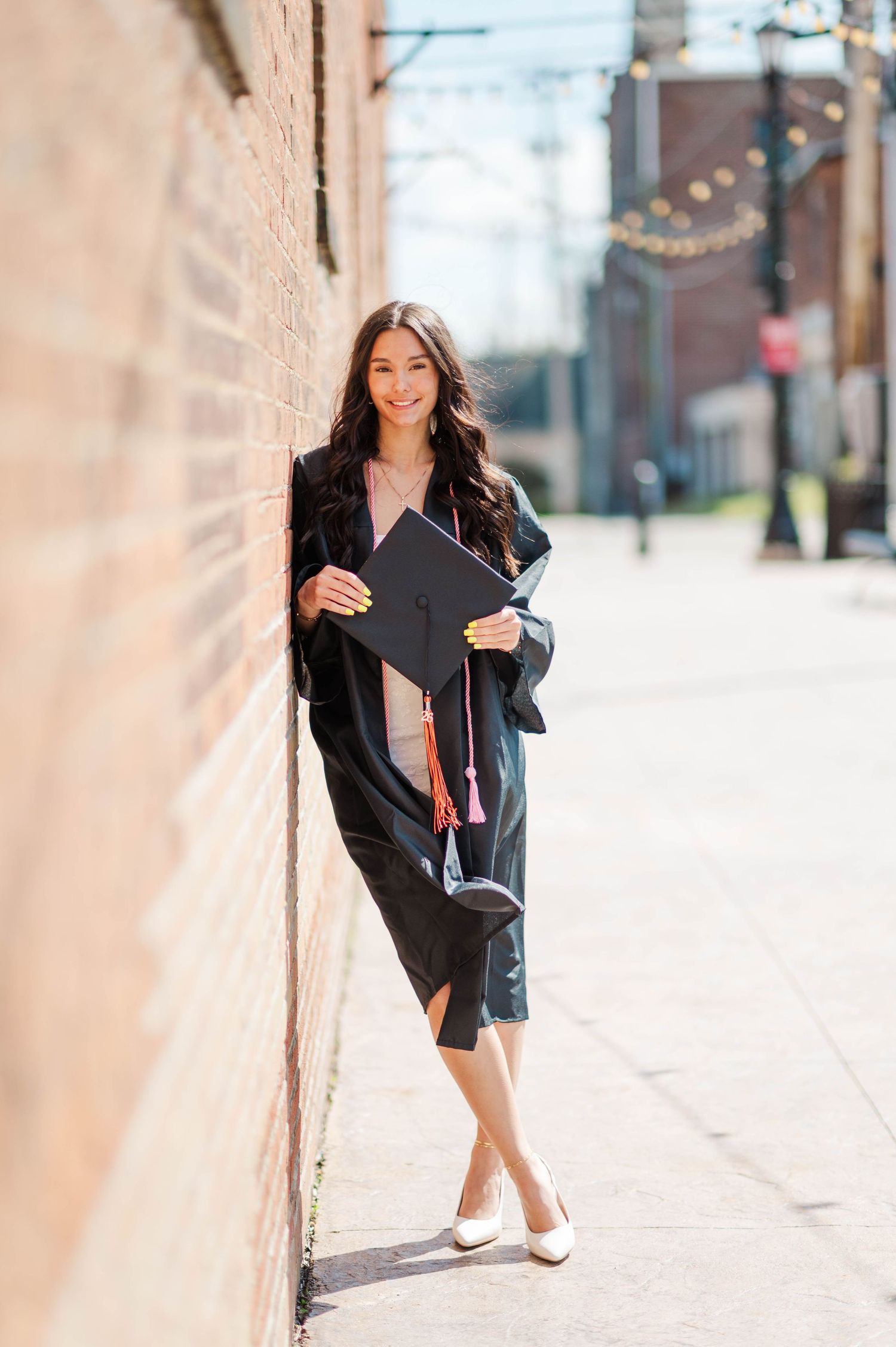 Smiling graduate in black gown holding cap with red tassel against brick wall outdoors.