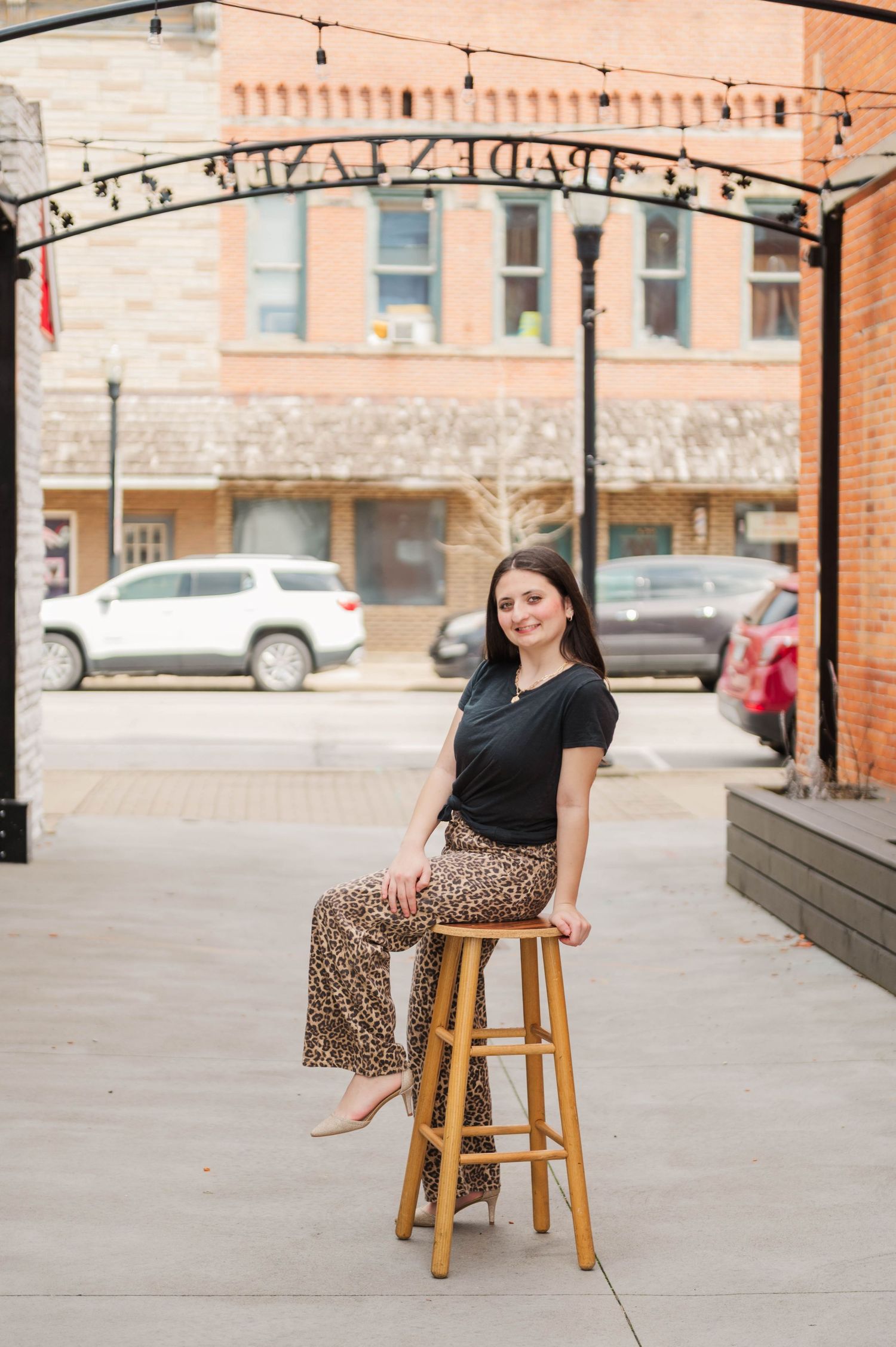 Young woman in leopard print pants sitting on a wooden stool under a decorative arch outdoors.