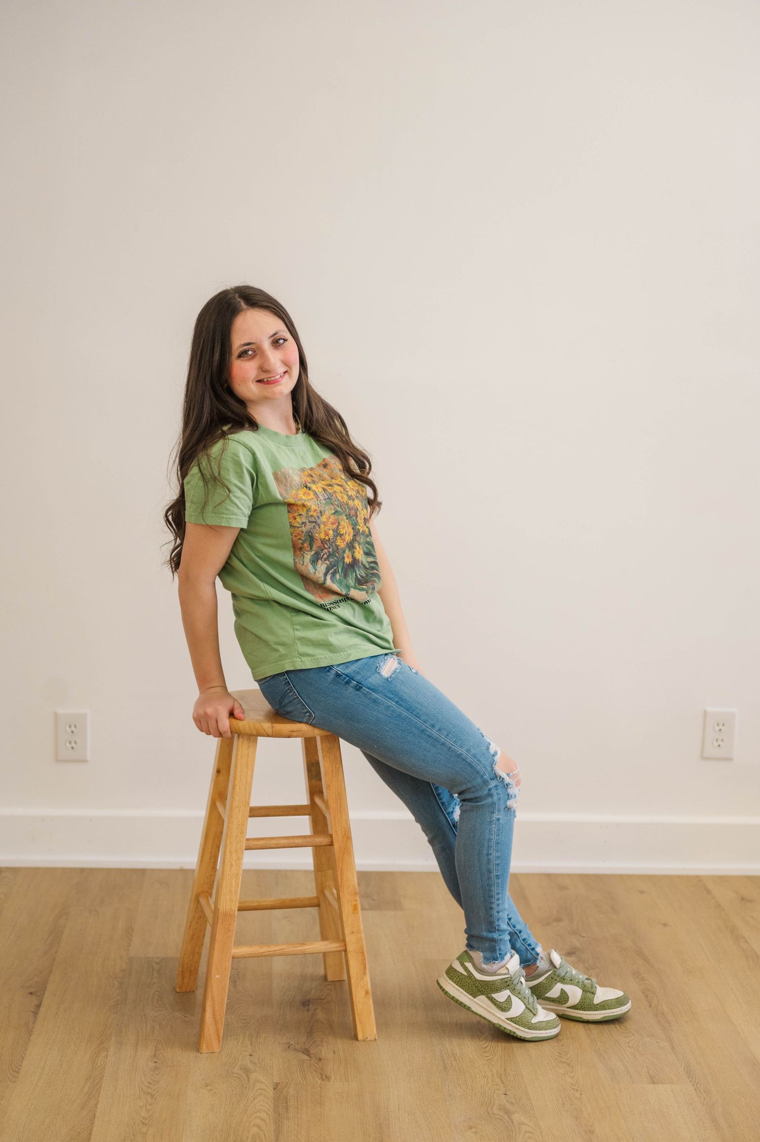 Brunette woman sits on wooden stool in green graphic tee, ripped jeans, and green sneakers.