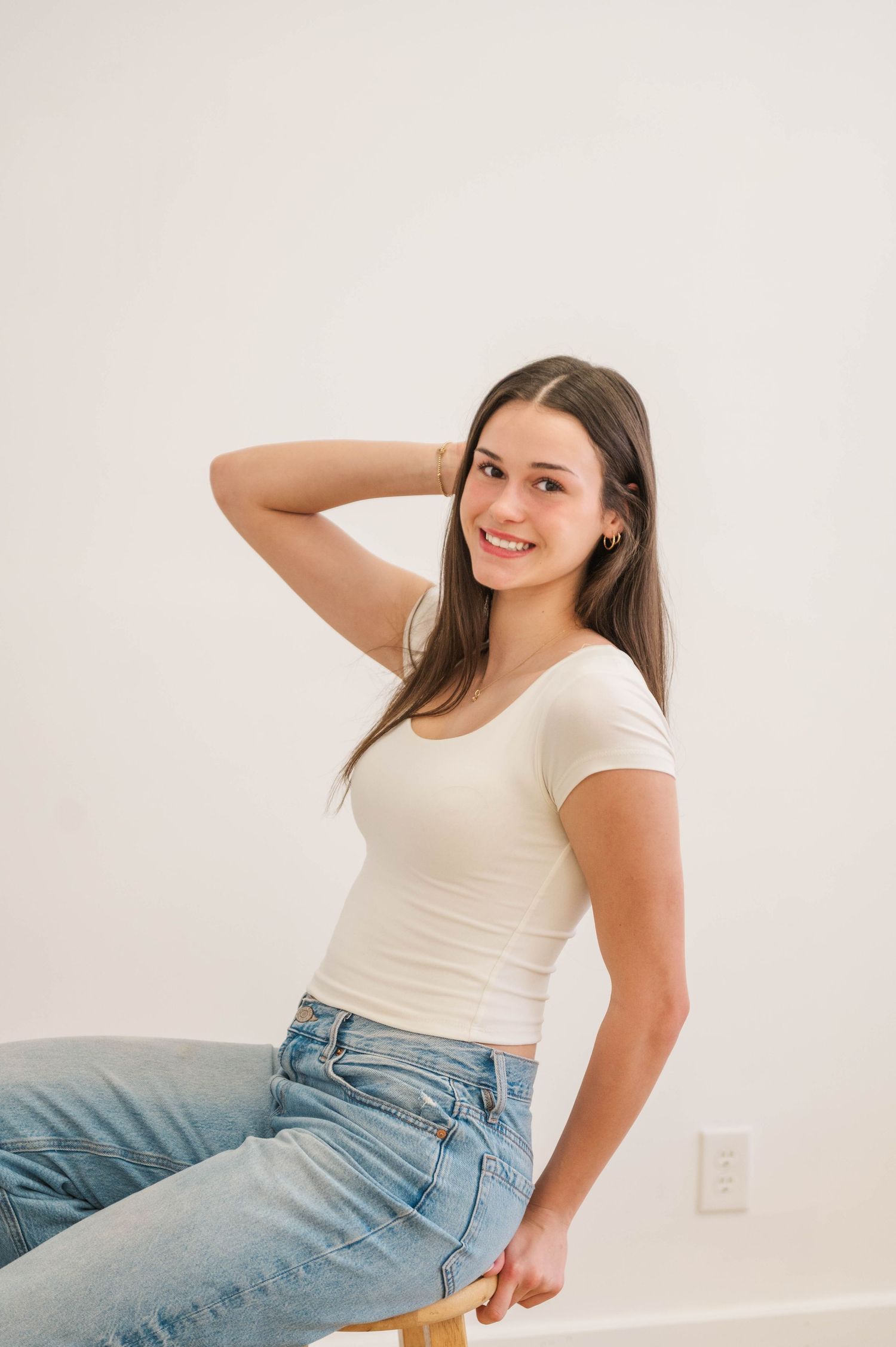 Smiling young woman with straight brown hair sits on stool in white tee and jeans, hand behind head.