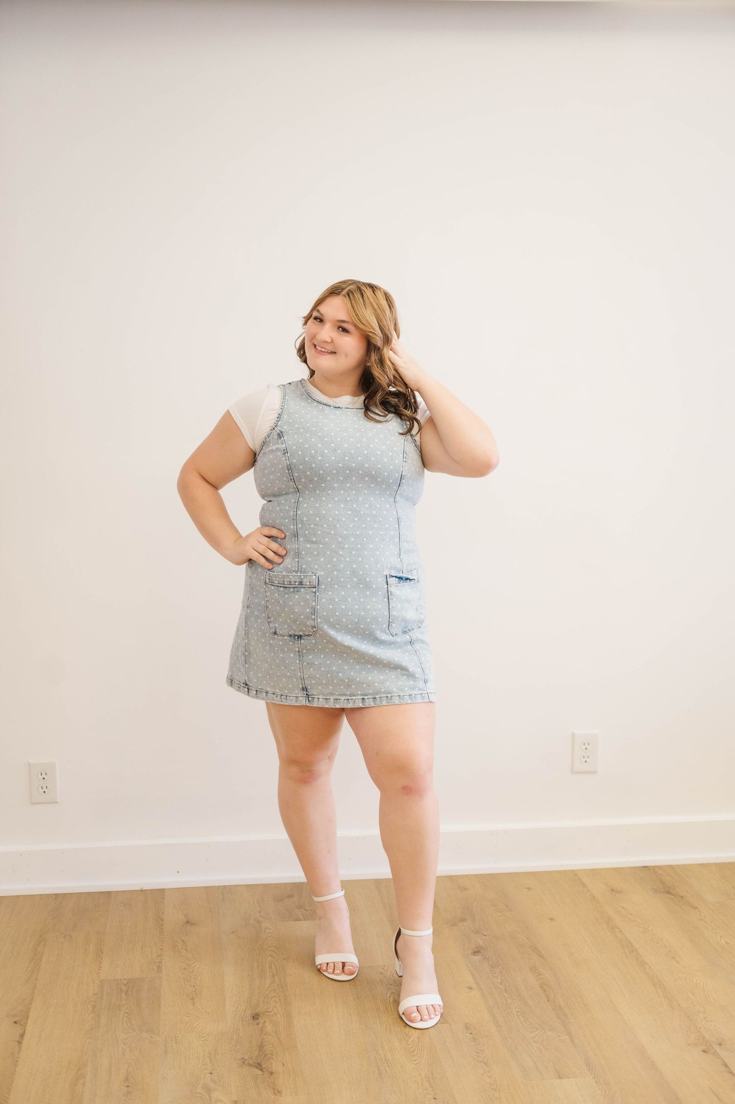Smiling woman in denim pinafore dress and white heels standing confidently against white wall.