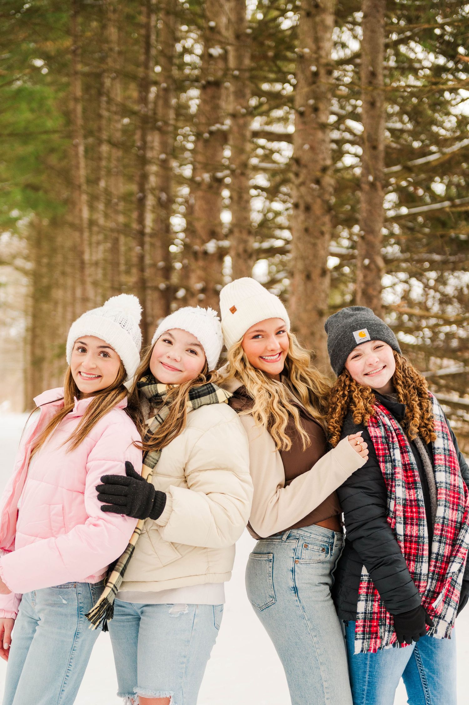 Four friends in winter coats and beanies pose together in a snowy forest path.