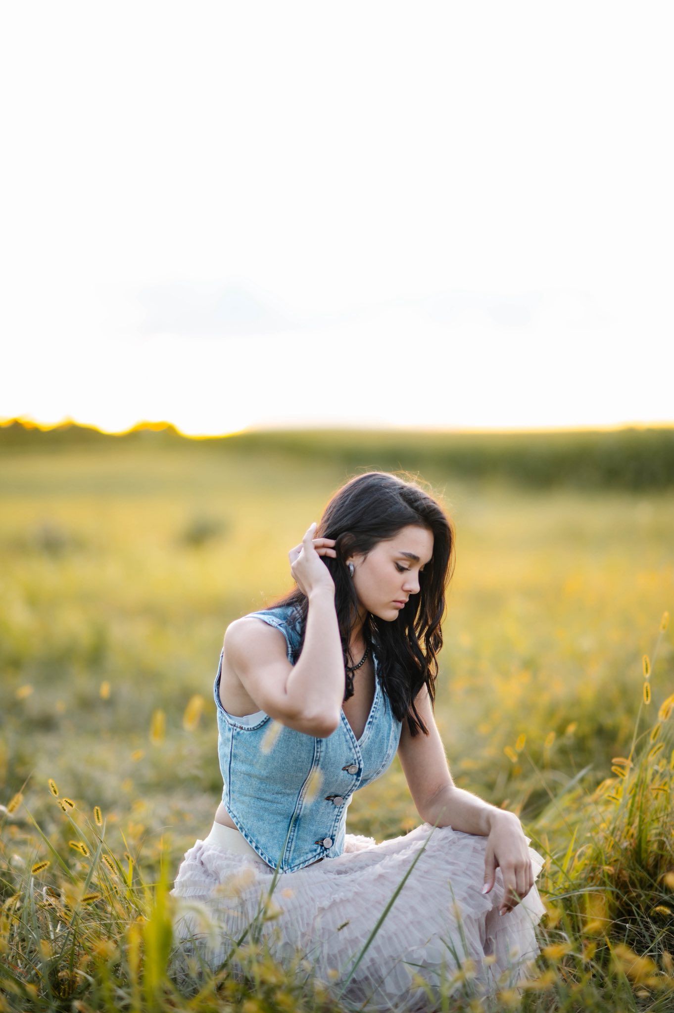 Artistic portrait of someone in a denim dress sitting thoughtfully among wildflowers at golden hour.