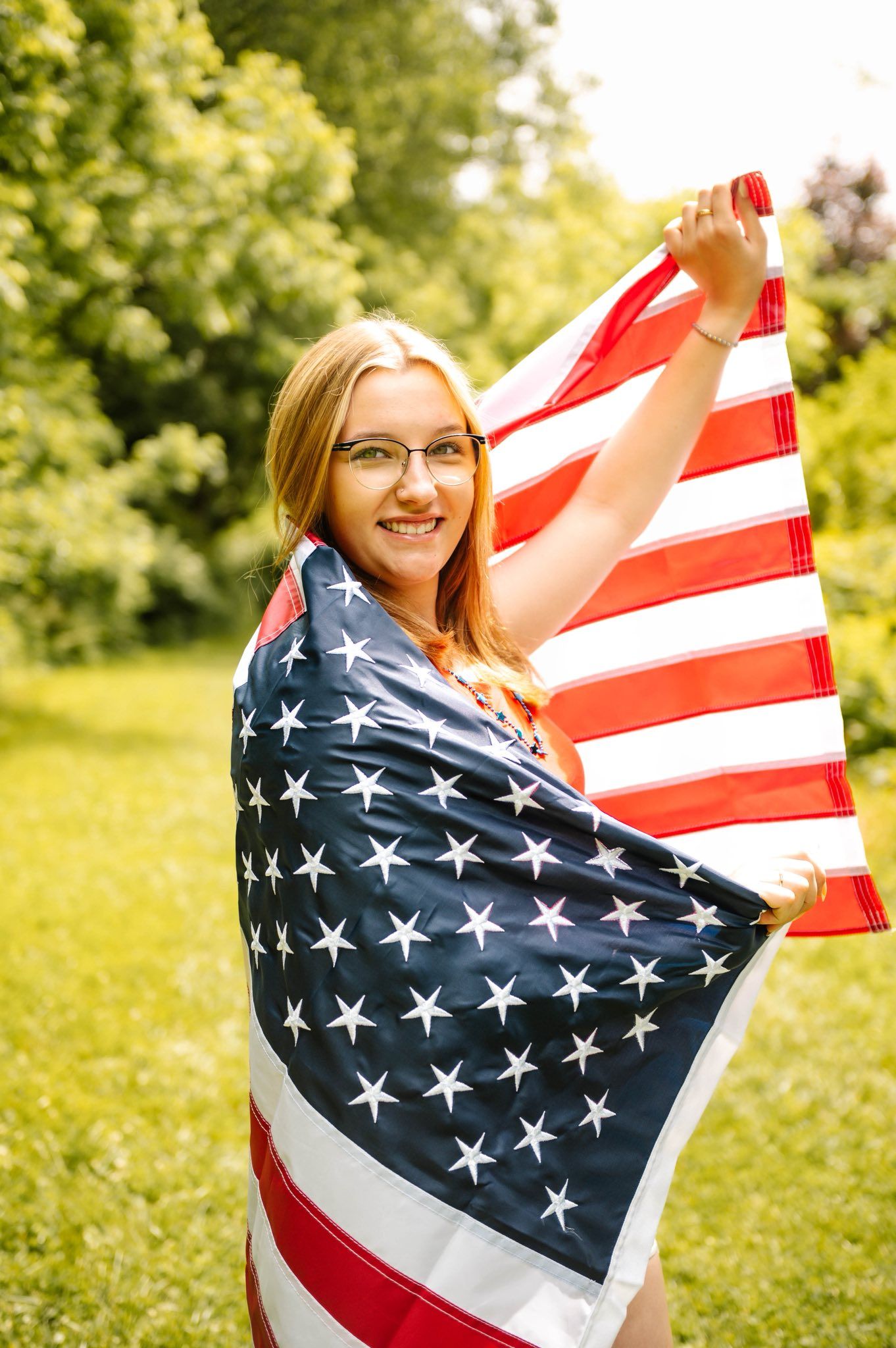 Person wrapped in American flag poses against sunny green background.