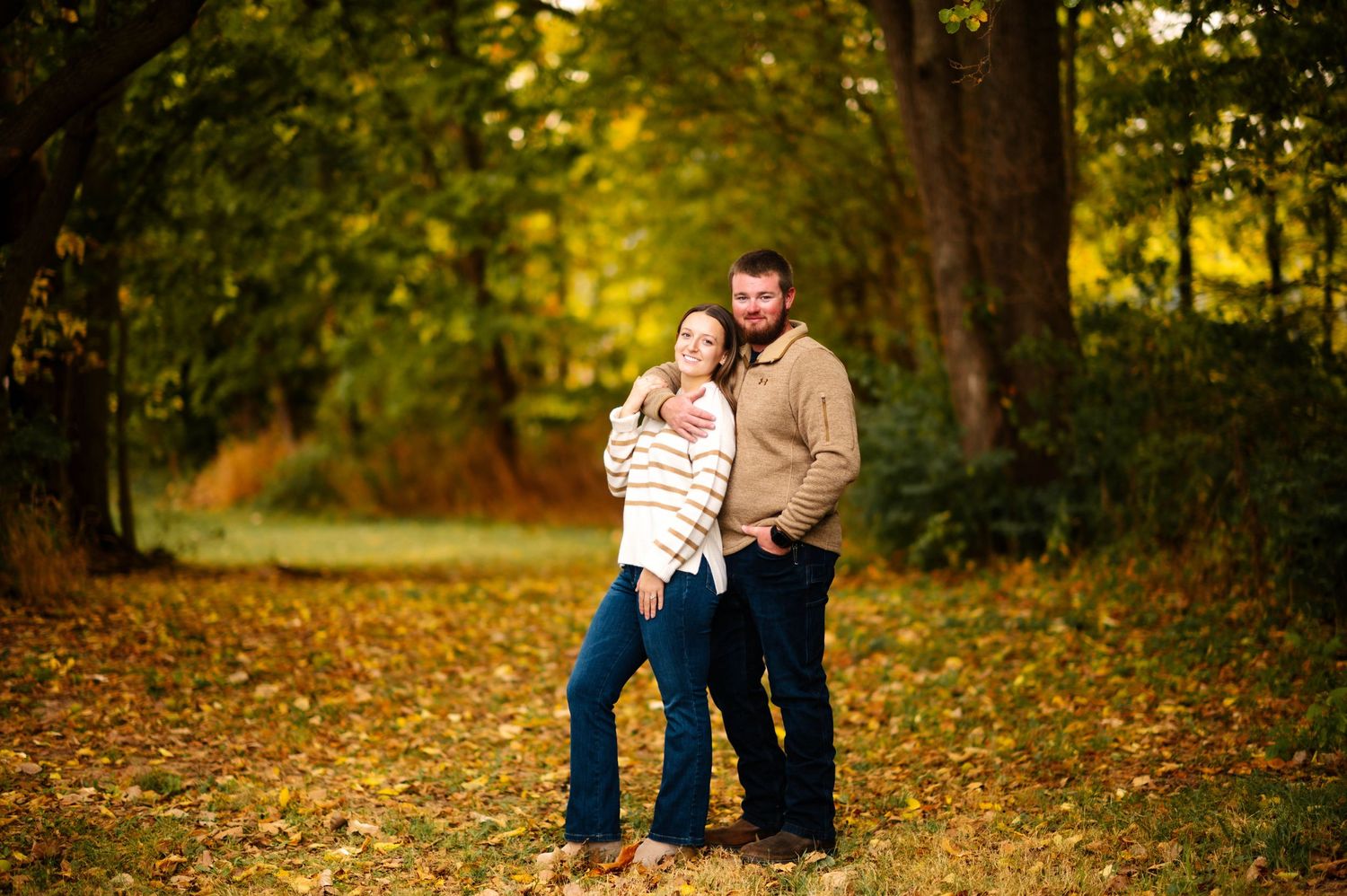 A couple poses together on fallen leaves in an autumn park setting.