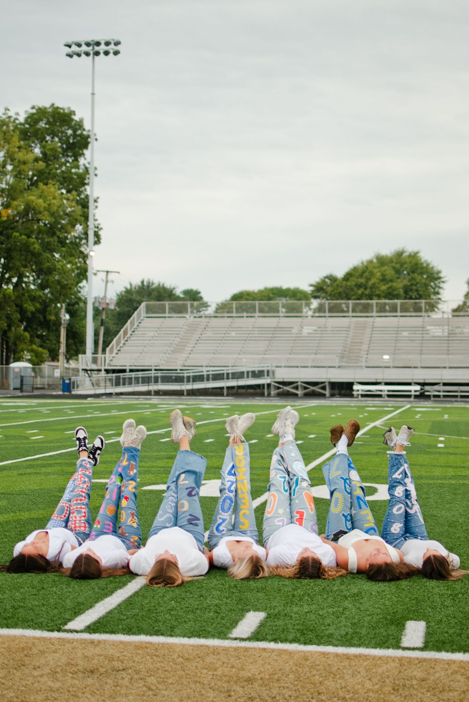 Six students lie on a football field with decorated senior jeans and legs raised in the air, with bleachers in the background.